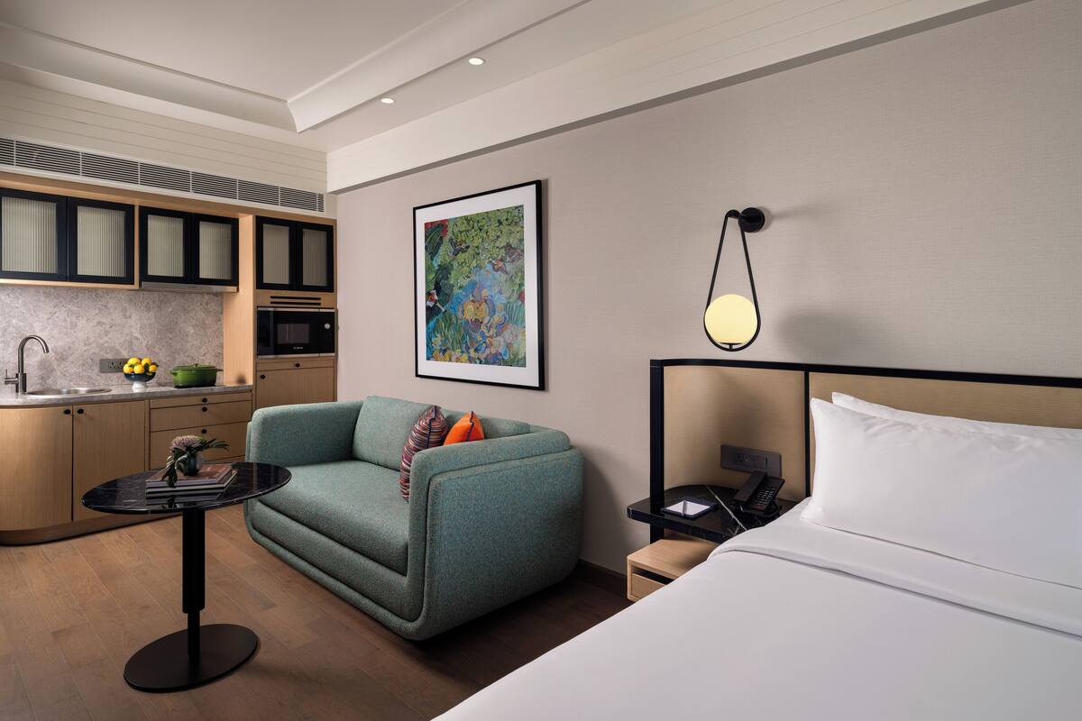 Una habitación de hotel moderna con sofá, una pequeña mesa, cama, mesita de noche, microondas y un cuadro en la pared.