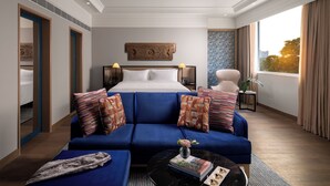 A modern hotel room with a large bed, a blue sofa, a small coffee table, and a view of the city through the window. - Pullman Chennai Anna Salai - Premium Brand By Accor (Chennai)