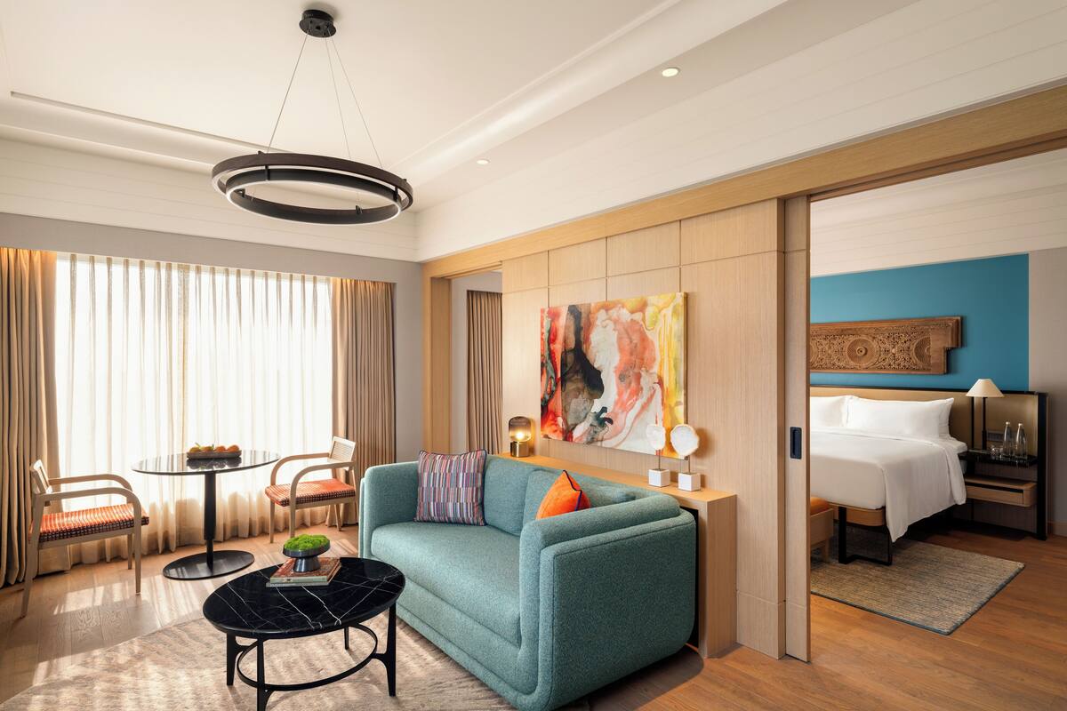 Una habitación de hotel moderna con un sofá verde azulado, una mesa de centro redonda negra, una zona de comedor con una mesita y sillas, y un cuadro abstracto grande en la pared.