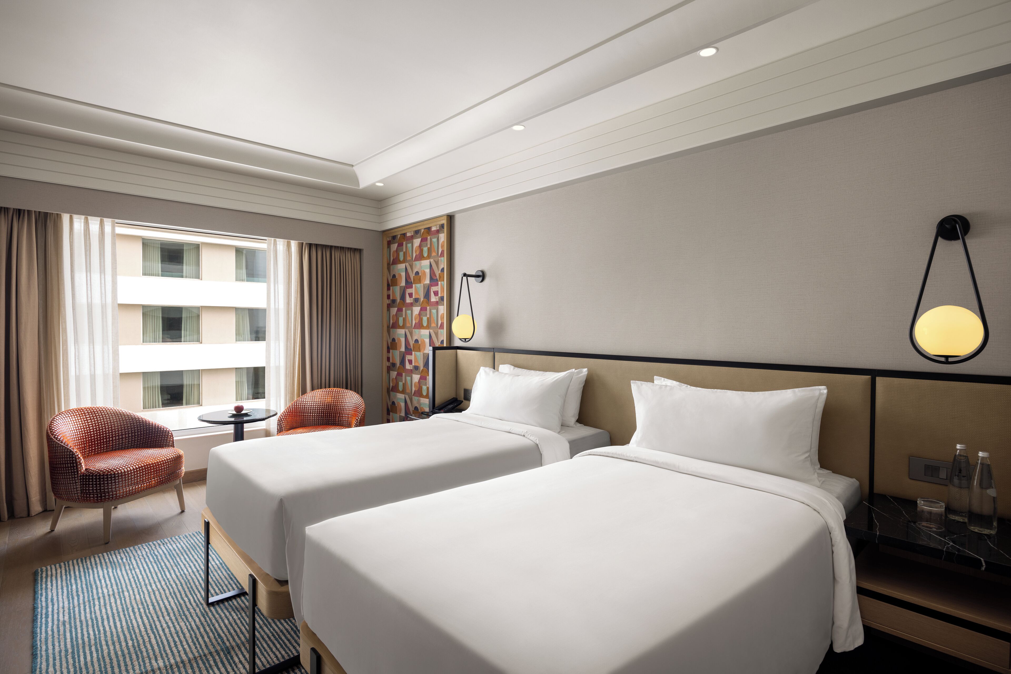 A hotel room with two beds, a small table, a chair, and a view of a building through the window.