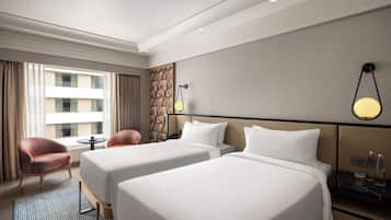 A hotel room with two beds, a small table, a chair, and a view of a building through the window.