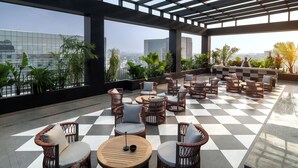 A rooftop terrace with wicker furniture, a checkered floor, and large windows offering city views. - Pullman Chennai Anna Salai - Premium Brand By Accor (Chennai)