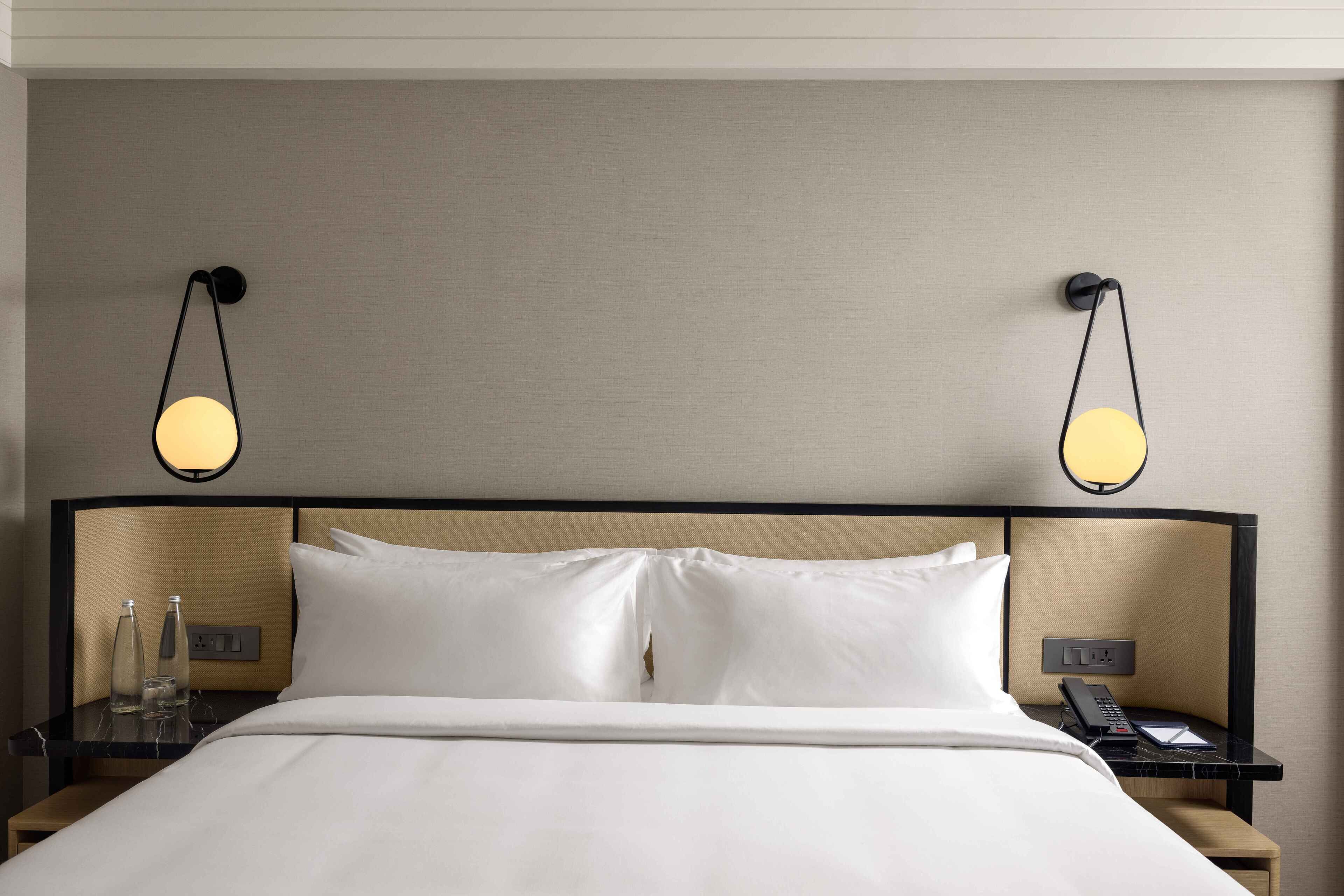 A neatly made bed with white linens, a bedside table with a lamp, a phone, and a bottle on the nightstand.