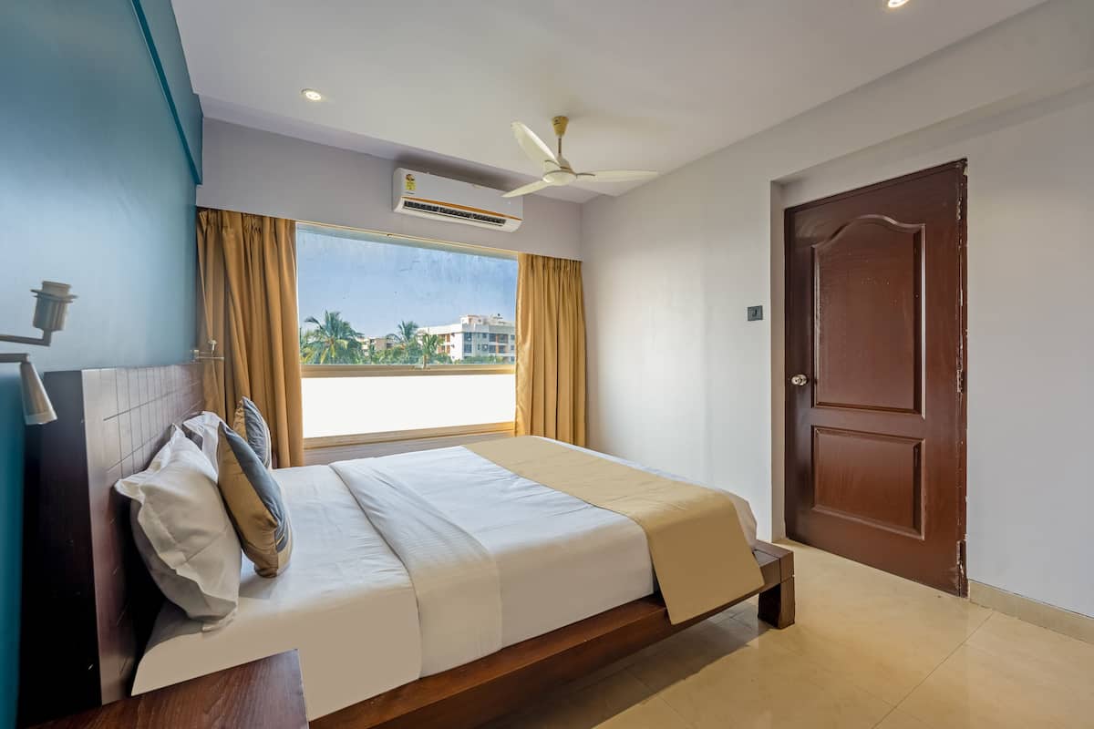 A hotel room with a bed, a ceiling fan, an air conditioning unit, a large window with curtains, and a wooden door.