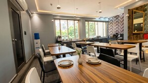 Un espace repas moderne, doté de plusieurs tables et chaises, de grandes fenêtres et d’une cuisine en arrière-plan.