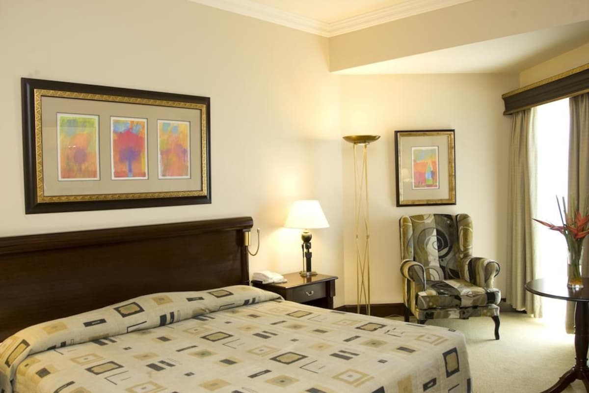 A hotel room with a bed, a chair, a nightstand, a lamp, and framed artwork on the wall.
