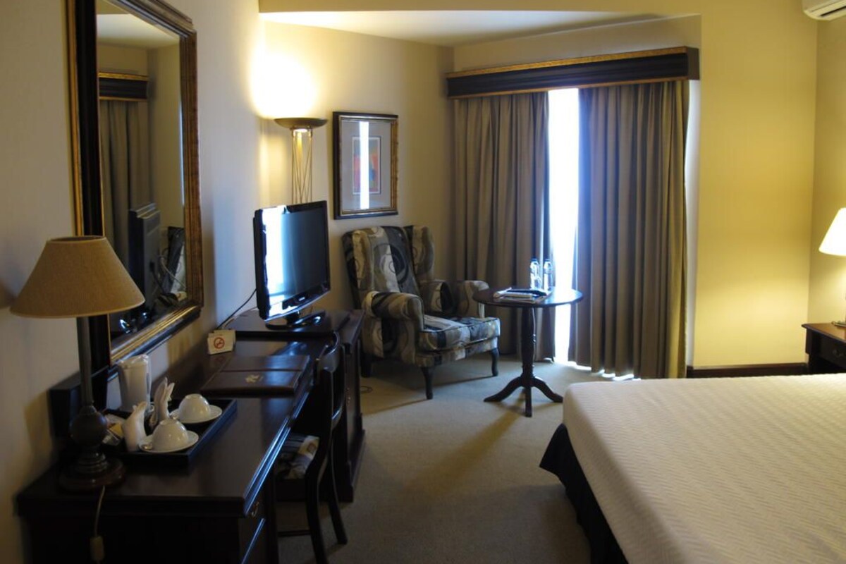 Une chambre d’hôtel avec un lit, un bureau avec une lampe, une chaise, une télévision et une fenêtre avec des rideaux.
