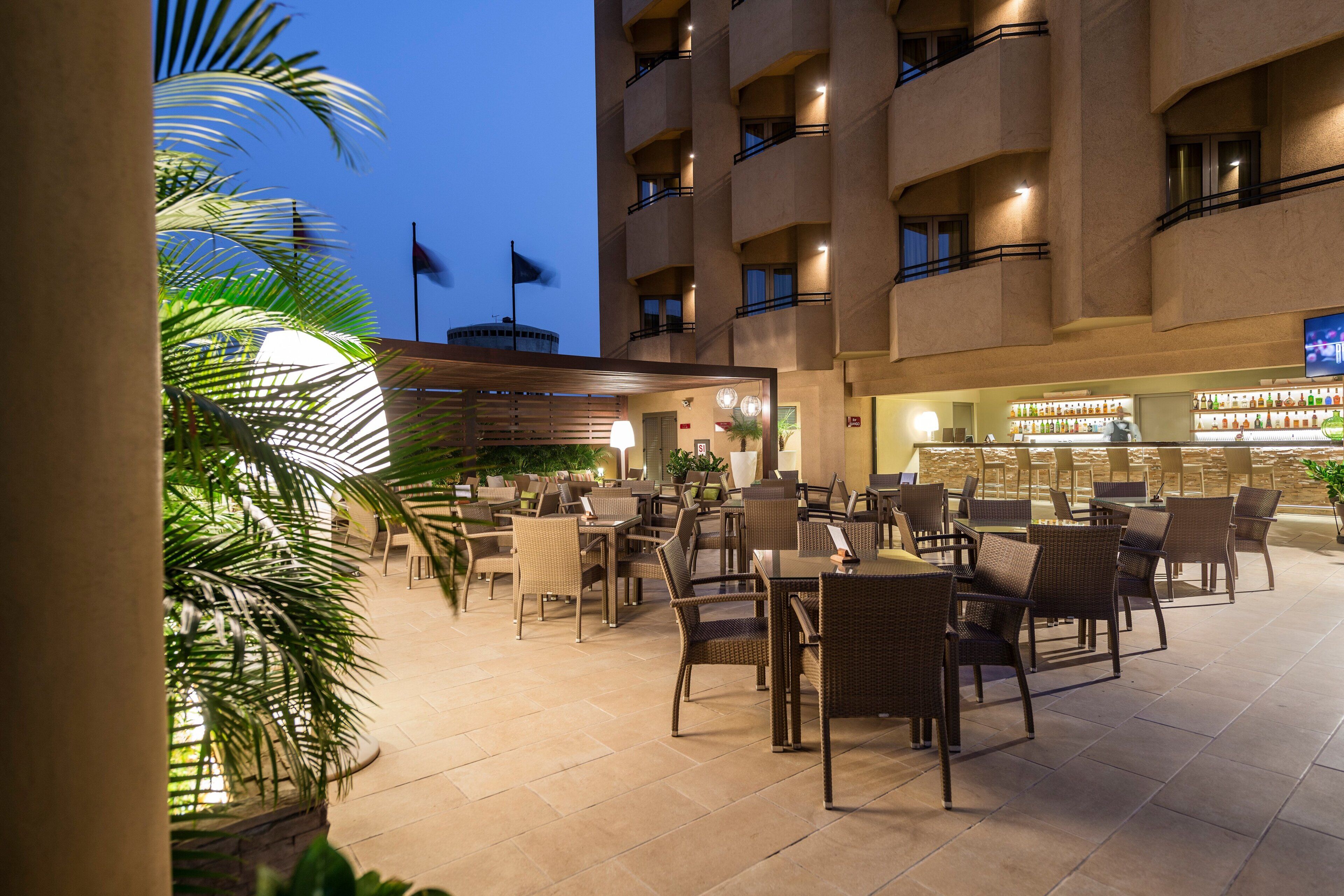 An outdoor dining area with wicker furniture and a tiled floor, surrounded by a building with multiple balconies and a flag on top.