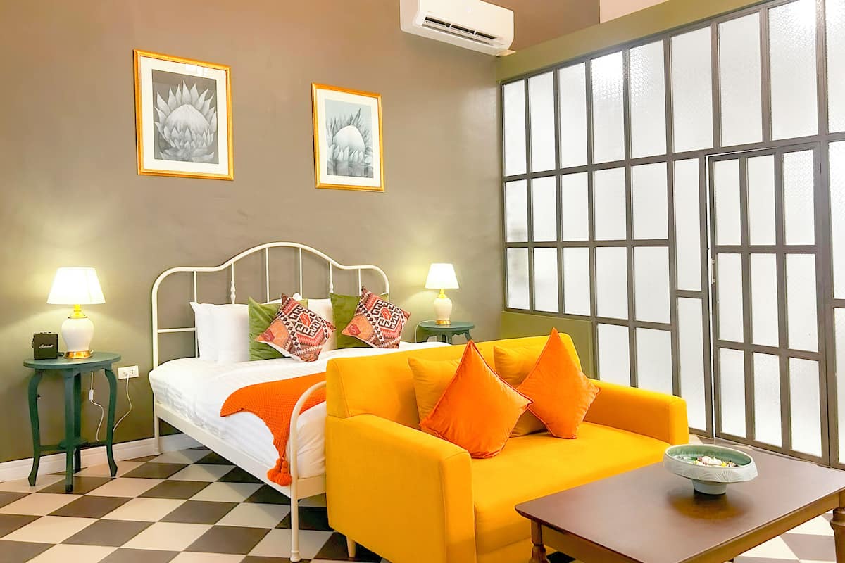 A hotel room with a bed, a yellow sofa, two side tables with lamps, a window with a view, and two framed pictures on the wall.