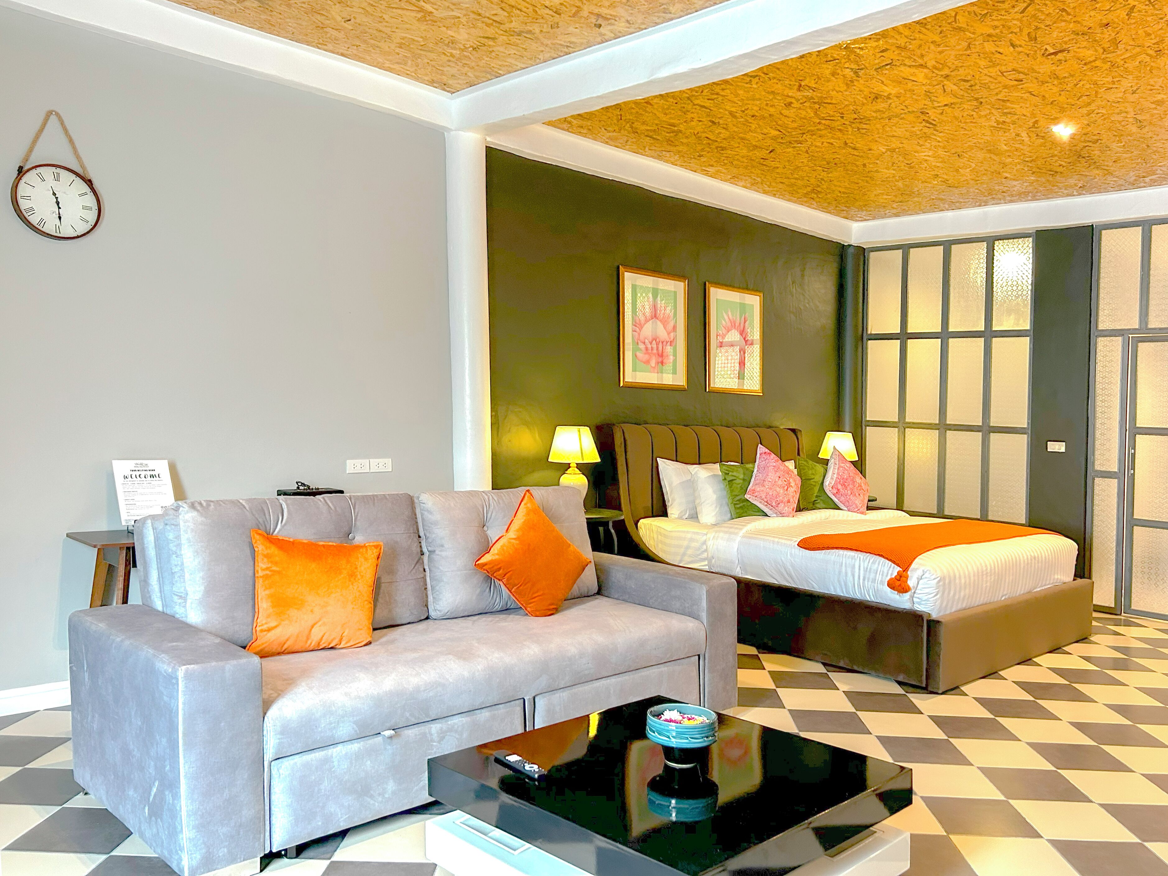 A hotel room with a grey sofa, a bed with orange bedding, a black coffee table, and a wall clock.