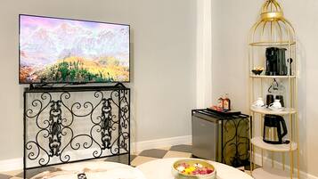 A living room with a TV, a coffee machine, a shelf with kitchenware, and a bowl of fruit on a marble table.