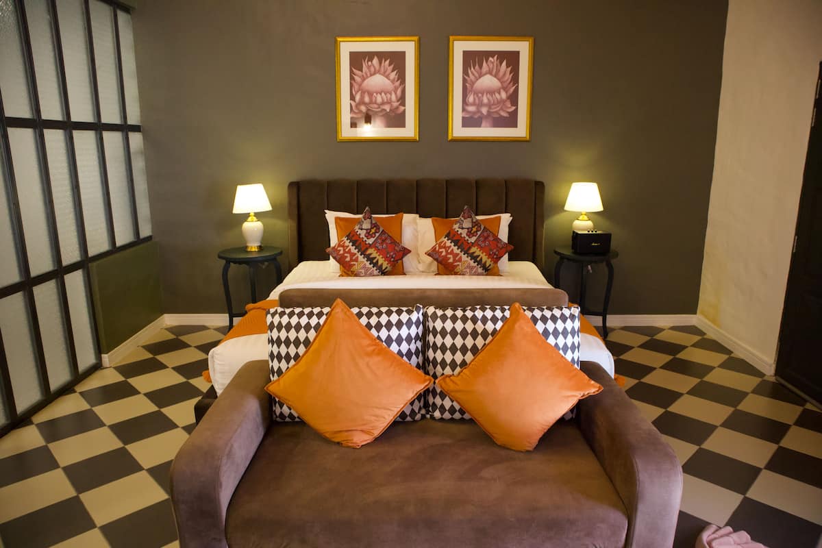 A hotel room with a checkered floor, a bed with patterned pillows, two wall-mounted lamps, and two framed floral pictures.
