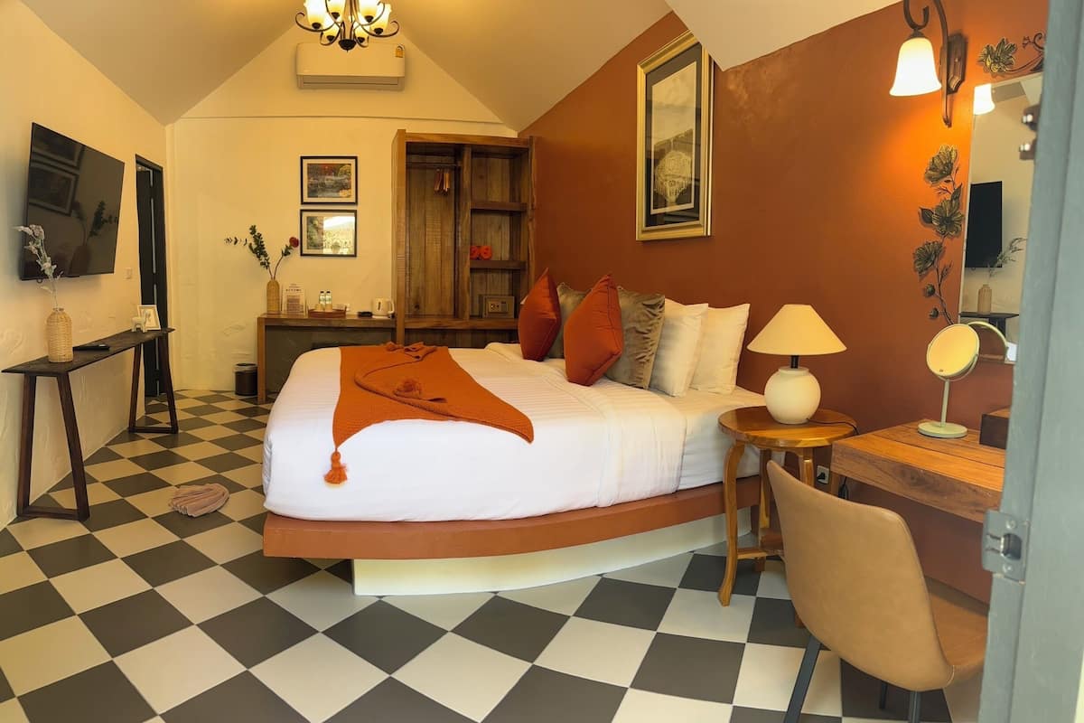 A hotel room with a checkered floor, a bed with a white comforter and orange throw, a wooden nightstand with a lamp, a desk with a mirror, and a wall-mounted TV.