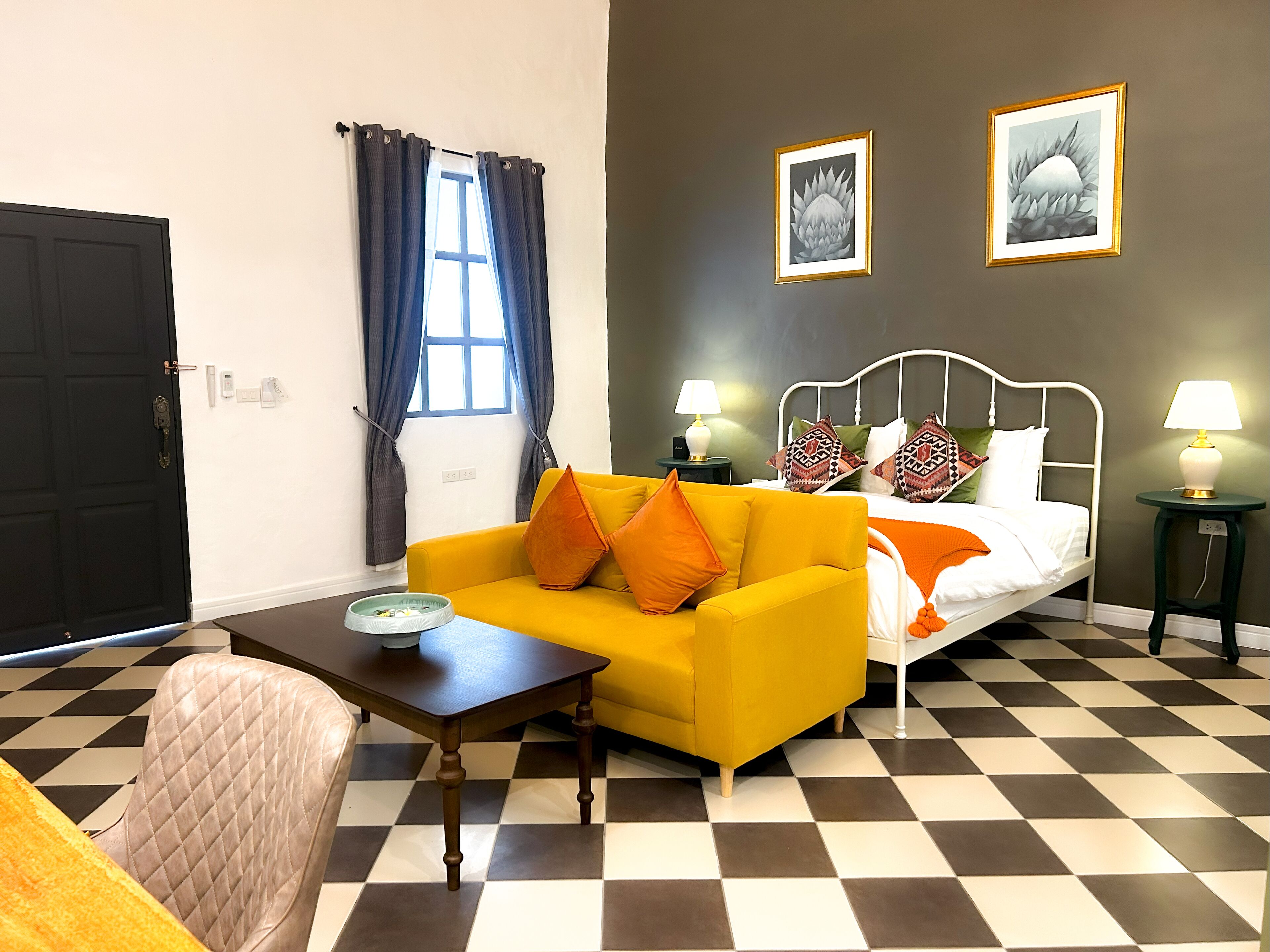 A room with a checkered floor, a yellow sofa with orange cushions, a white bed with patterned pillows, two bedside lamps, and two framed pictures on the wall.