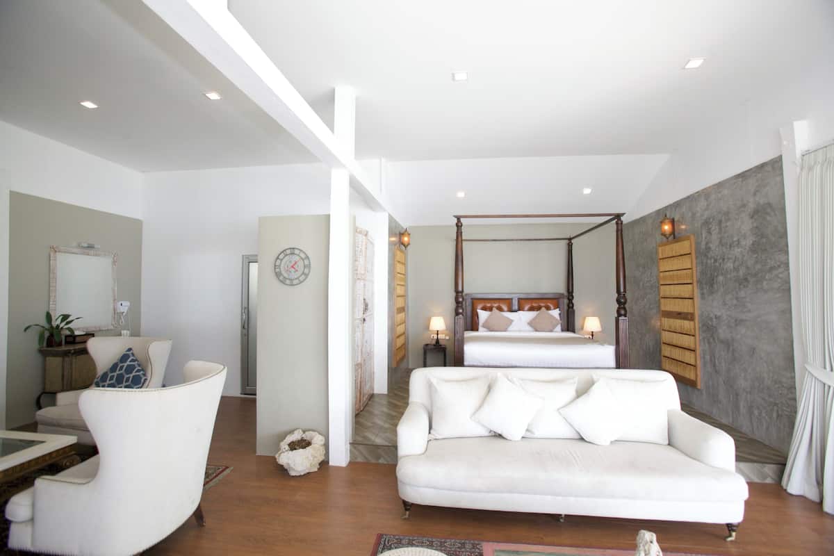 A modern hotel room with a large bed, a sitting area with a white sofa, a desk, and a large mirror.