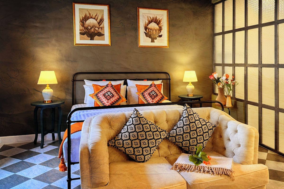 A hotel room with a checkered floor, a bed with patterned pillows, a sofa with decorative pillows, two wall-mounted lamps, and a large window with a view.