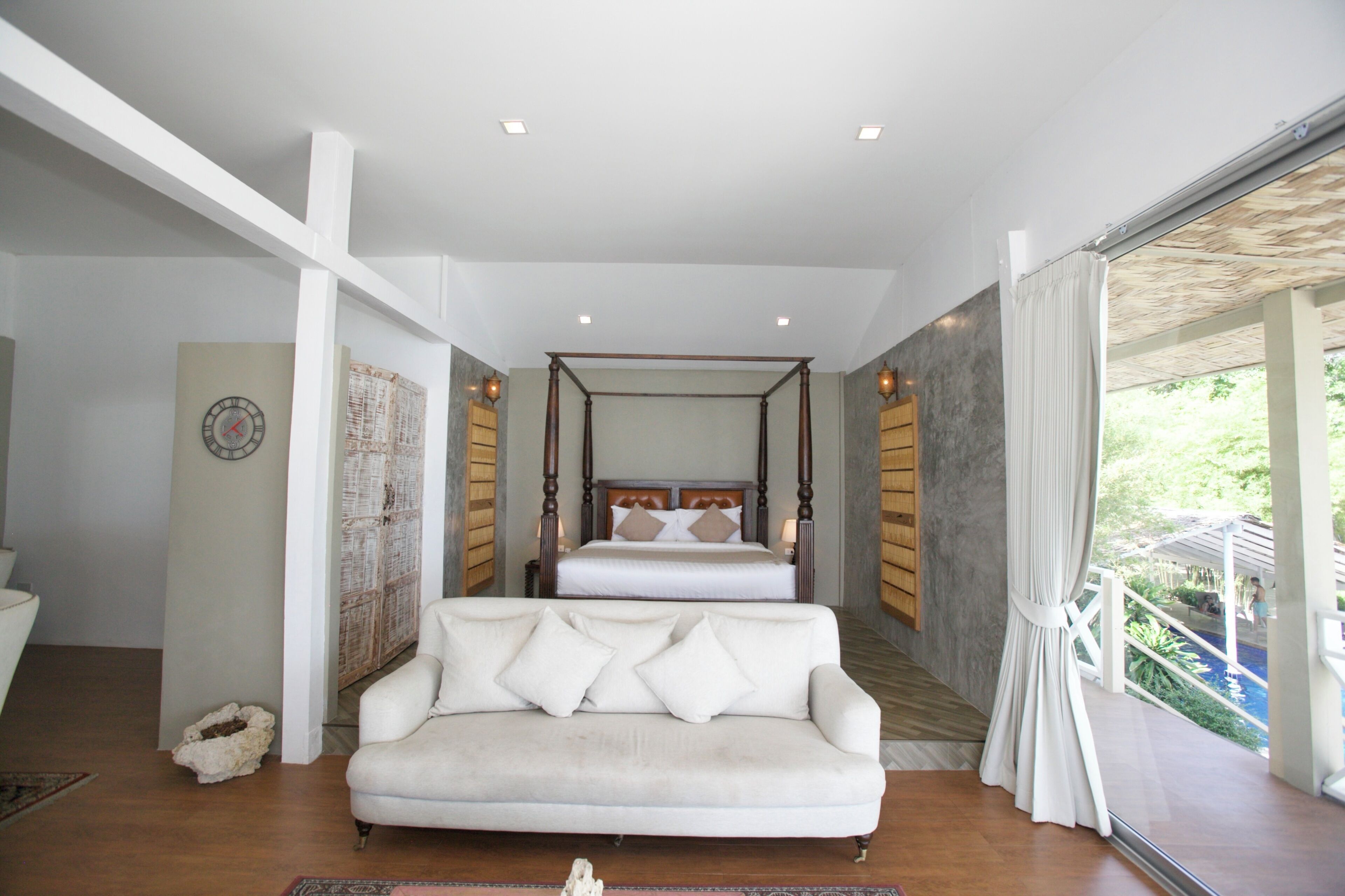 A bedroom with a four-poster bed, a white sofa, and a view of a pool.