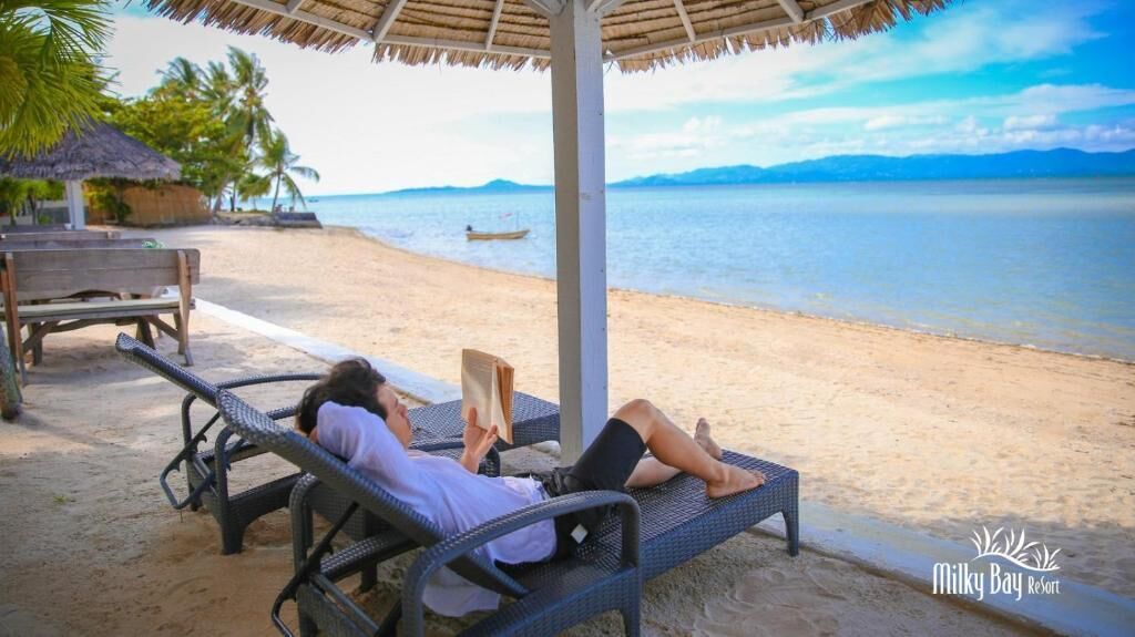 Escena de playa con una persona descansando en una silla de descanso bajo una techumbre de paja, un banco de madera y un bote pequeño en el agua.