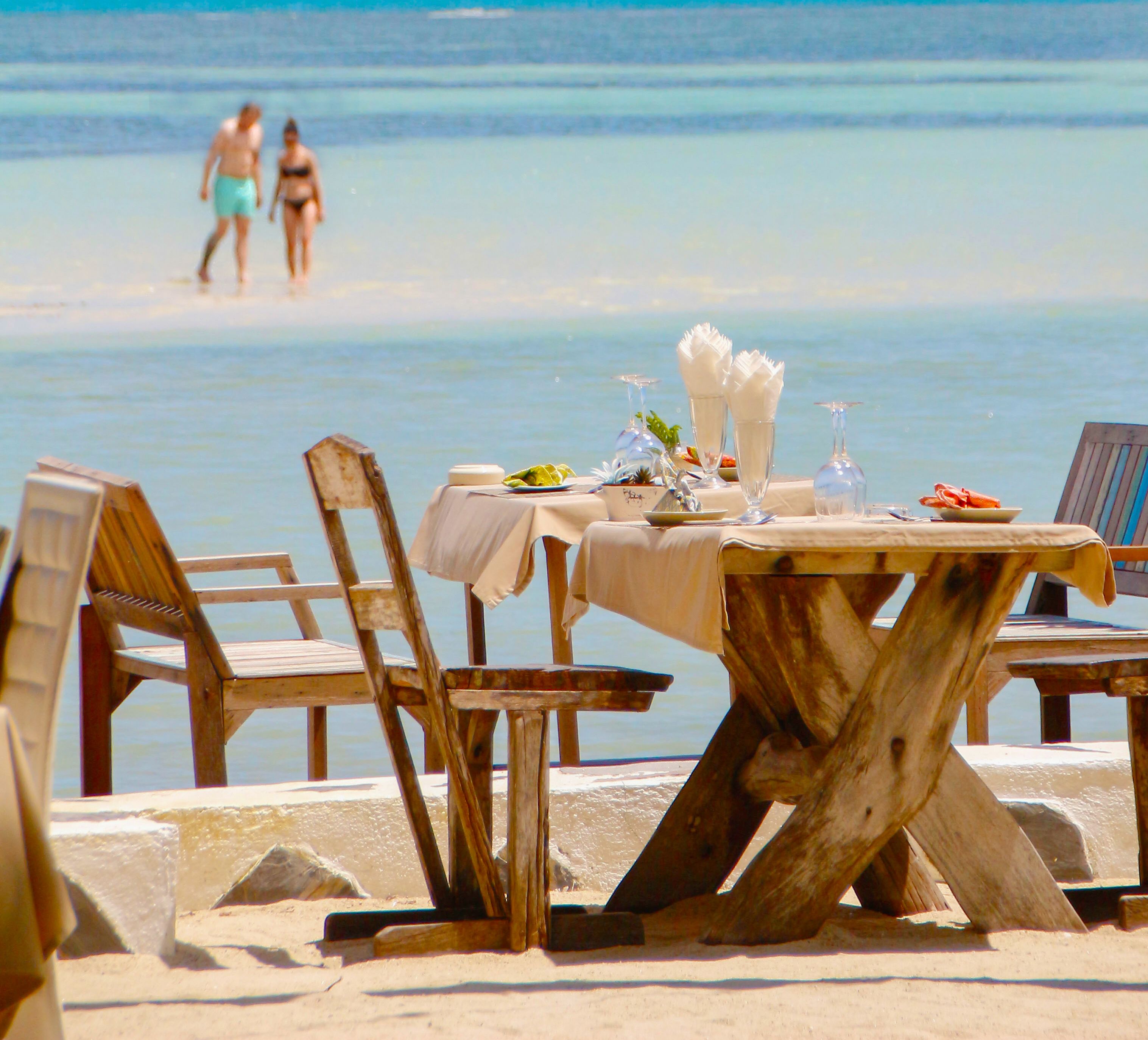 A beachside table set with a white tablecloth, wooden chairs, and a view of the ocean.