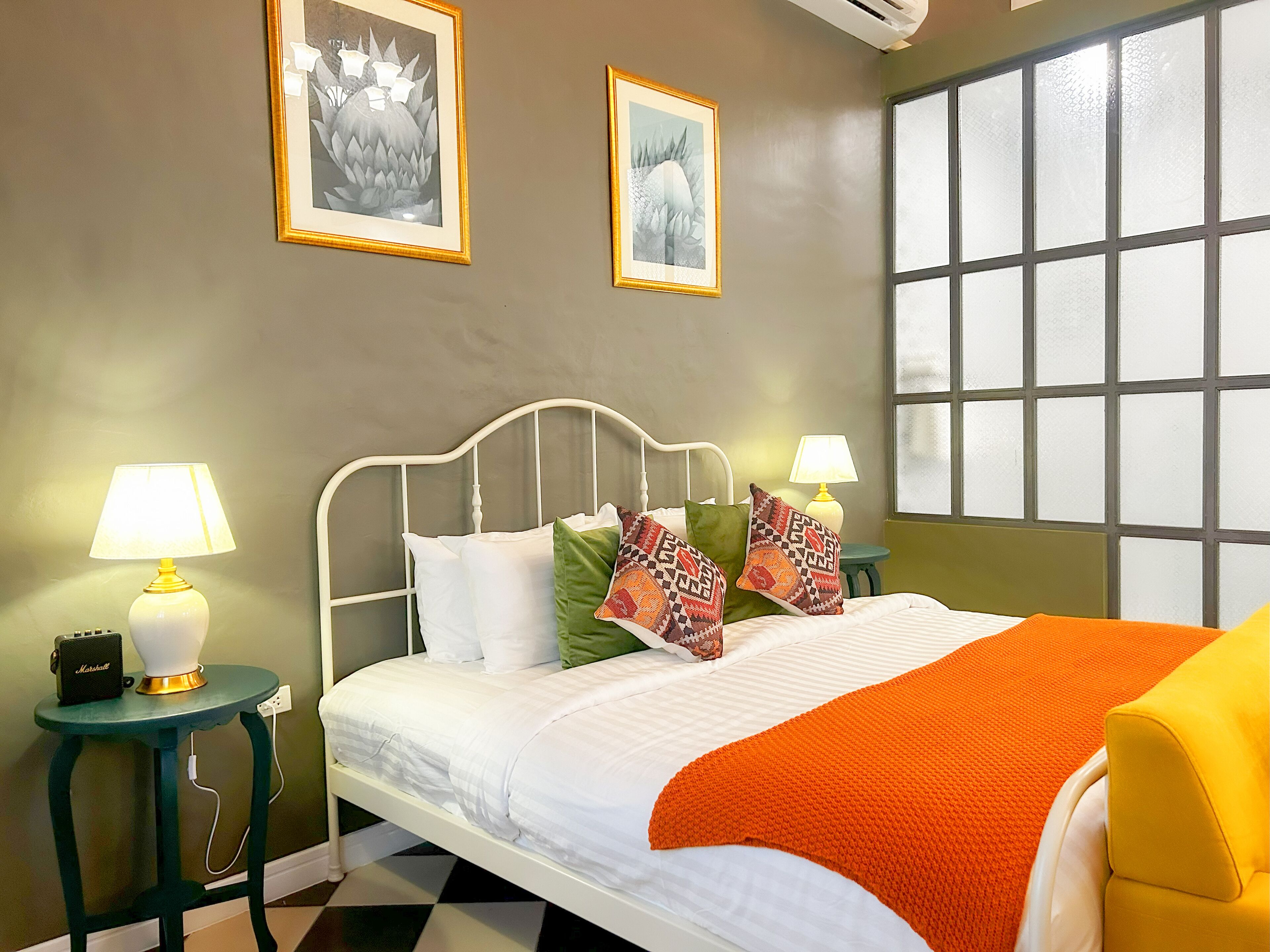 Een net opgemaakt bed met wit beddengoed en een oranje plaid, met aan beide kanten nachtkastjes en lampen, waarvan er één een Marshall speaker heeft.