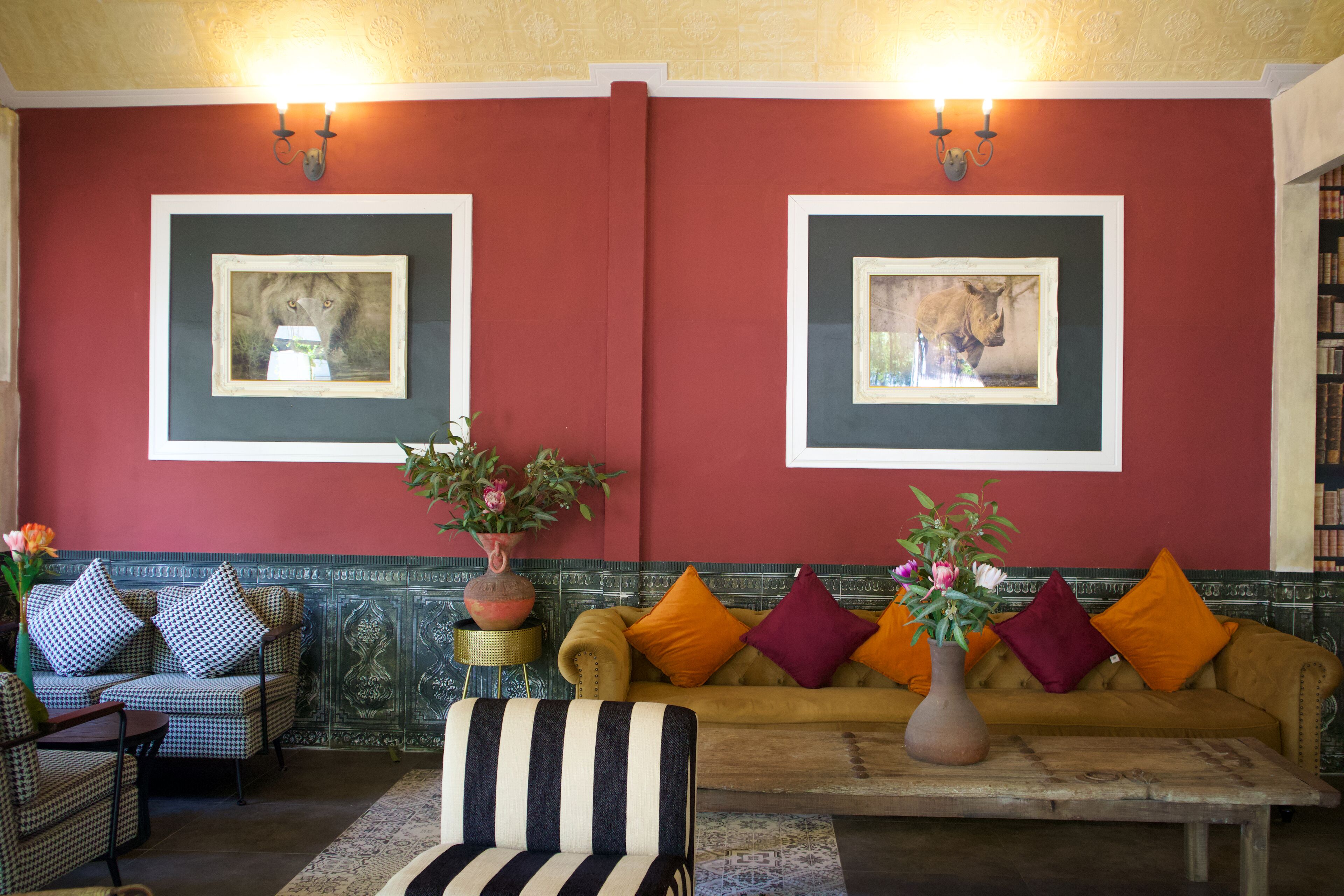 Una habitación con una pared roja, dos cuadros enmarcados de animales, un sofá con cojines naranja y morados, un sillón a rayas y una mesa con un jarrón de flores.