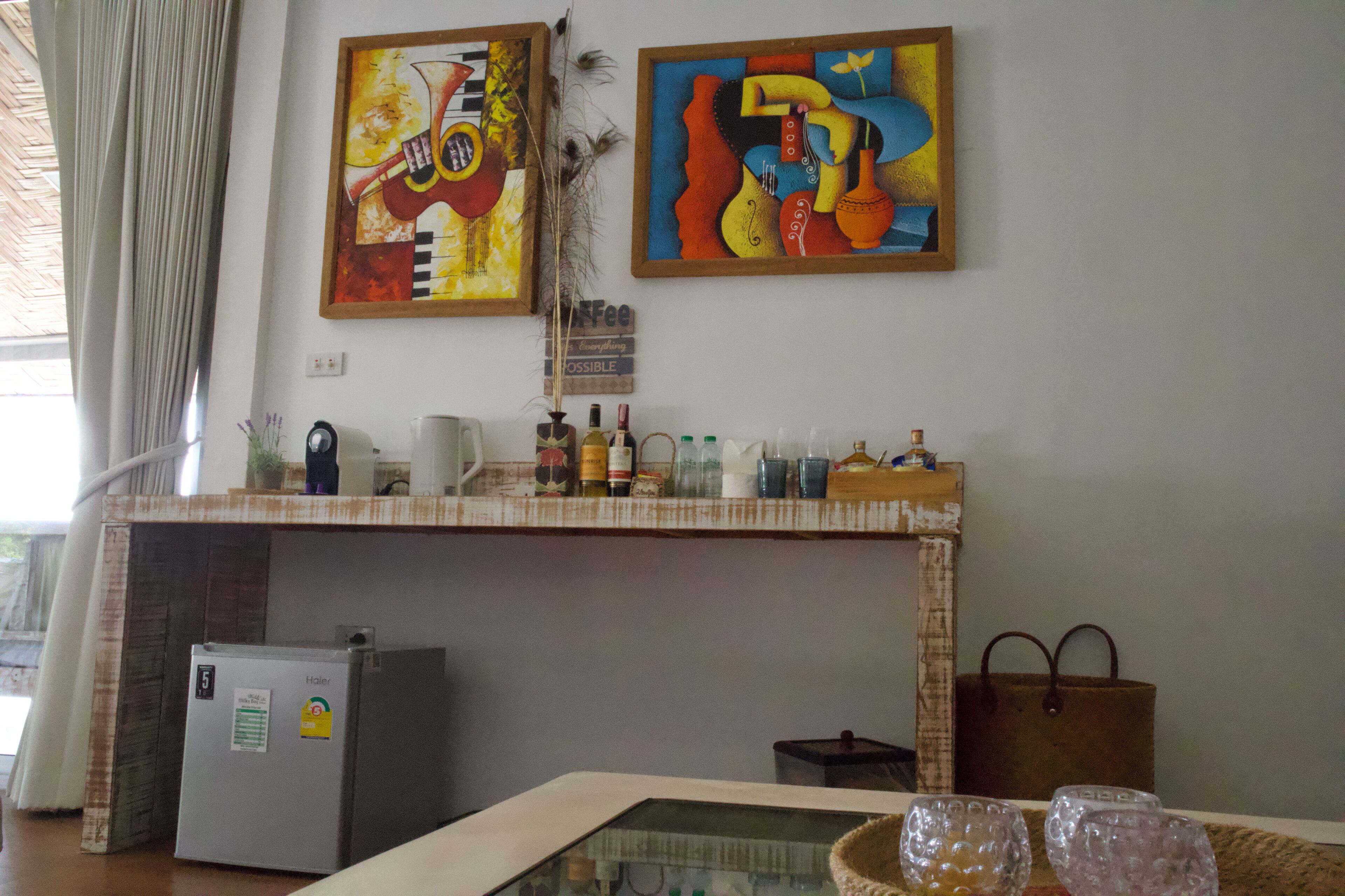 Een kamer met een kleine koelkast, een tafel met glazen blad en verschillende flessen en glazen, en twee schilderijen aan de muur.