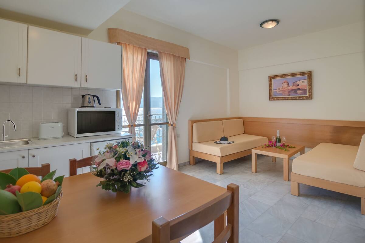 Eine moderne Küche mit weißen Schränken, einer Mikrowelle und einem Toaster. Im Essbereich steht ein Tisch mit einer Blumenvase und einer Obstschale. Im Wohnbereich befindet sich eine Couch und ein kleiner Tisch mit einer Flasche und Gläsern.