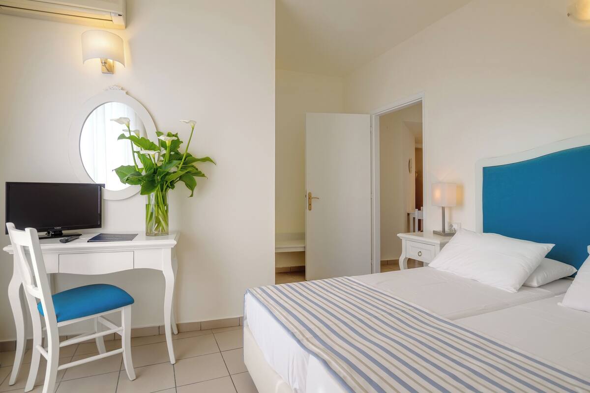 Ein Hotelzimmer mit einem Bett, einem Schreibtisch mit Computer, einem Stuhl, einer Pflanze und einem Spiegel.