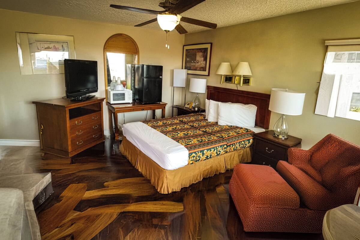 A hotel room with a bed, a desk with a television, a microwave, a nightstand with lamps, and a red chair.