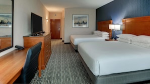 A hotel room with a desk, two beds, a television, a lamp, and a cityscape painting on the wall.