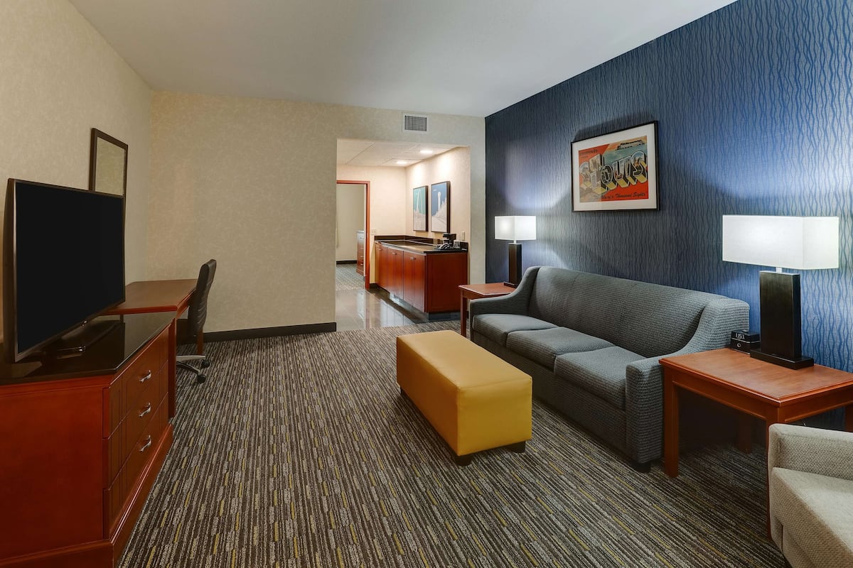 Habitación de hotel con televisor, sofá, una mesita, lámpara y suelo alfombrado.