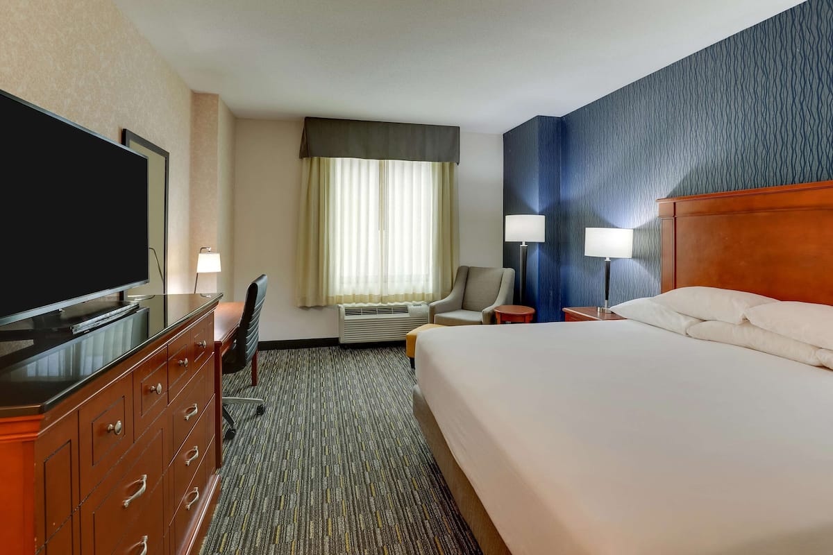 Una habitación de hotel con una cama grande, televisión, escritorio con silla, lámpara y ventana con cortinas.