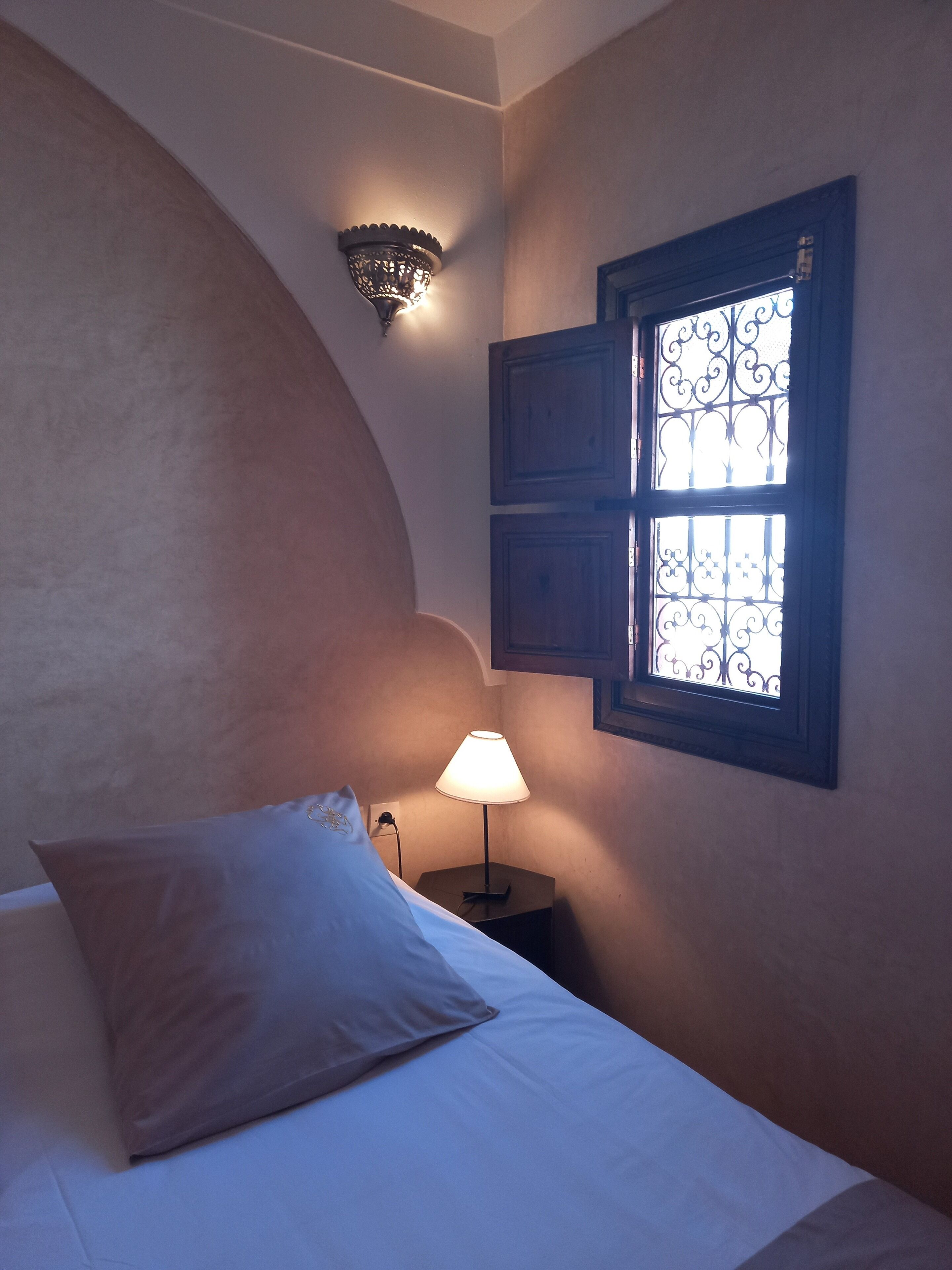 A bedroom with a bed, a nightstand, a wall-mounted lamp, and a window with decorative iron bars.