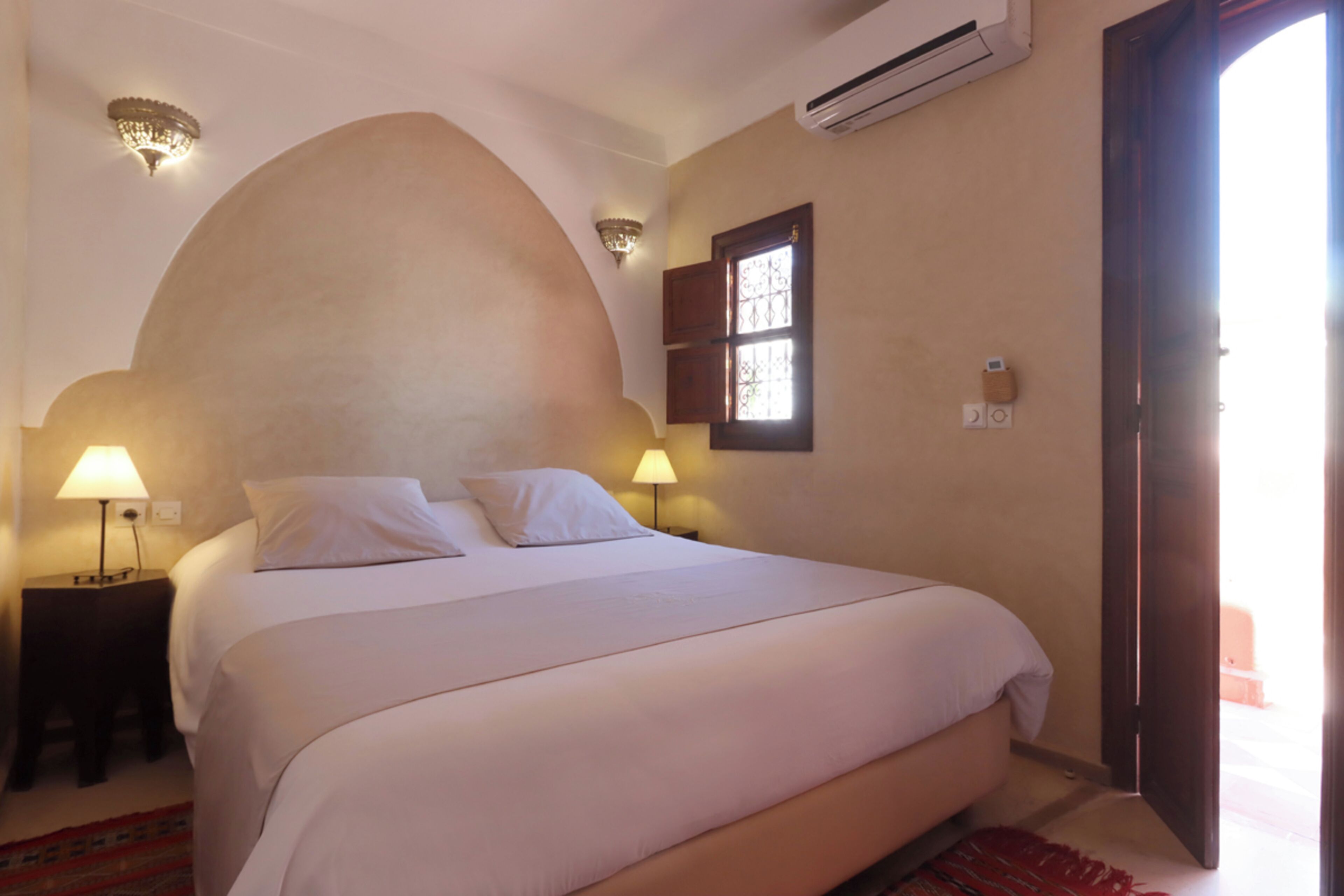 A hotel room with a large bed, two bedside lamps, an air conditioning unit, a window with a view, and a door.