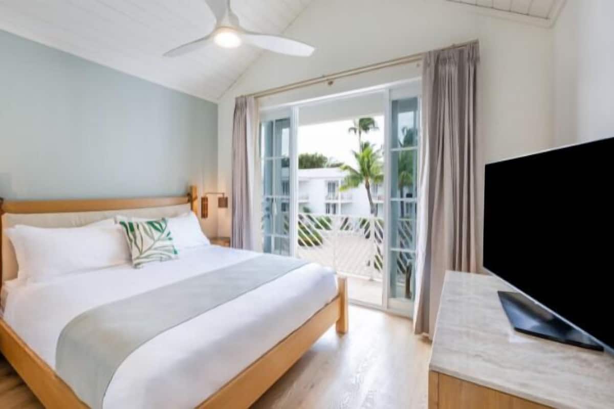 A hotel room with a bed, a ceiling fan, a TV, and a balcony with a view of palm trees.