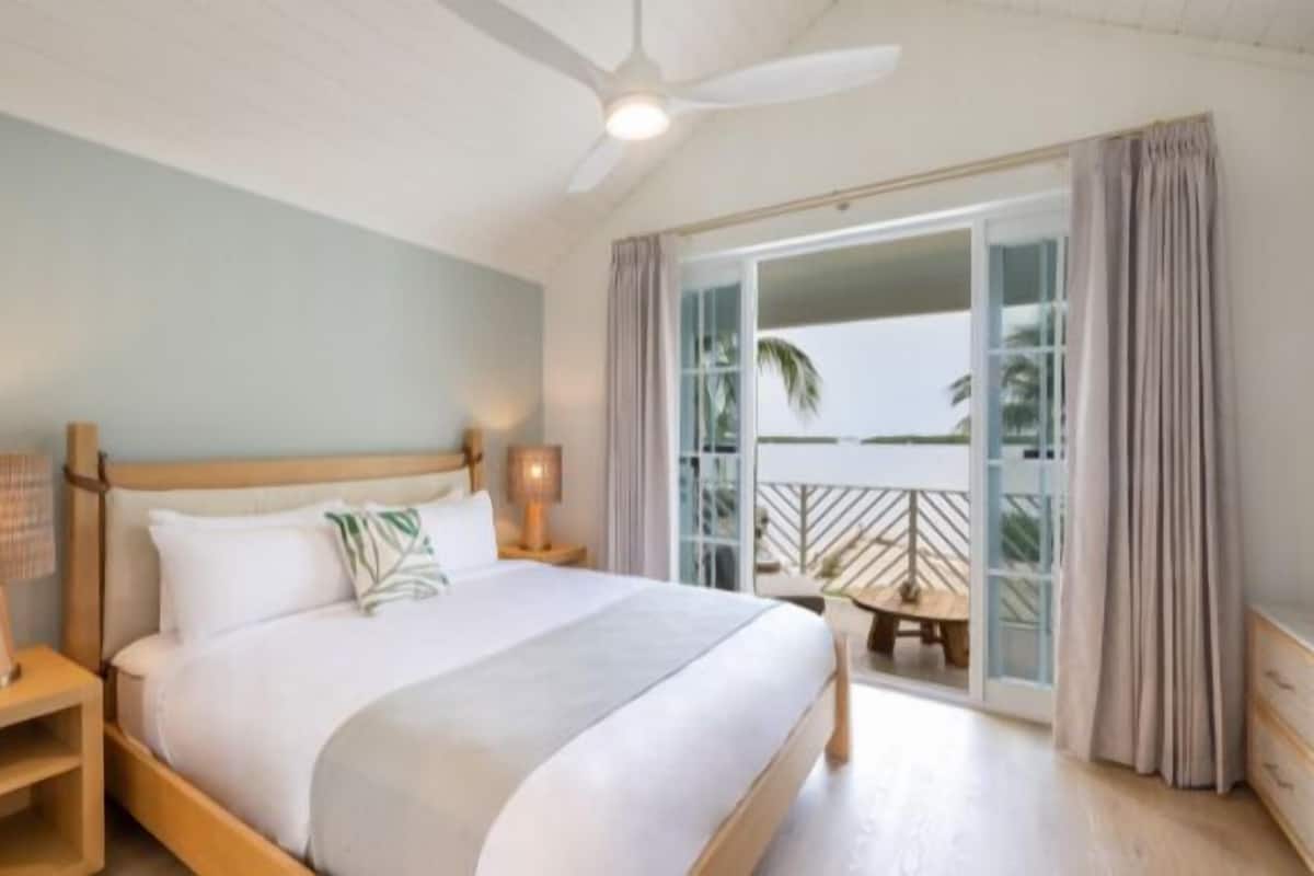 A bedroom with a bed, bedside lamps, and a view of the beach through a sliding glass door.