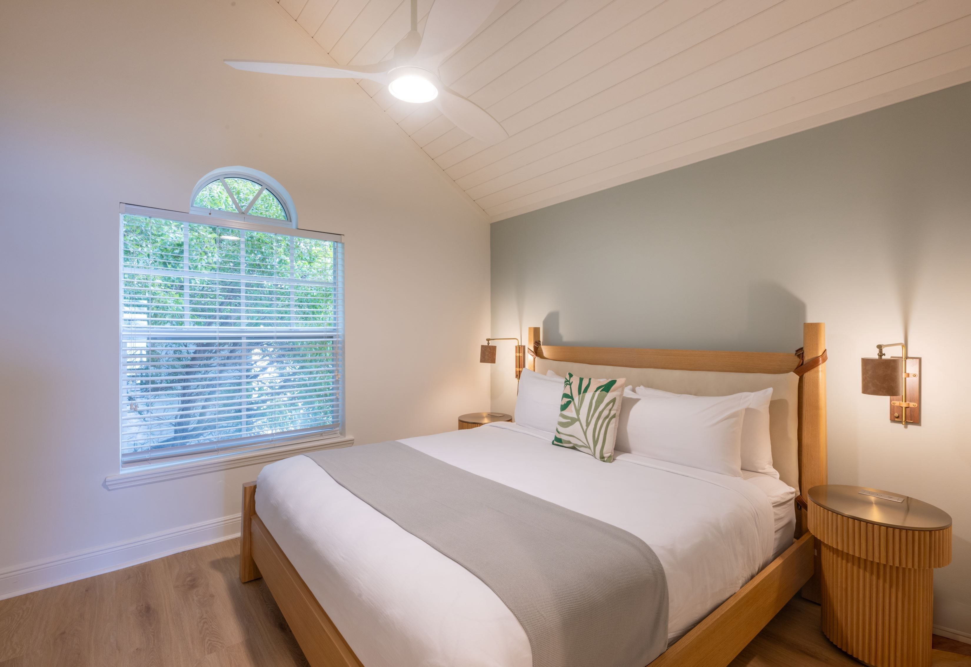A bedroom with a large bed, a ceiling fan, a window with blinds, and a nightstand with a lamp.