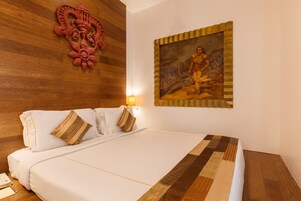 A neatly made bed with white linens and decorative pillows, a wooden headboard, a wall-mounted decorative piece, and a framed painting above the bed.
