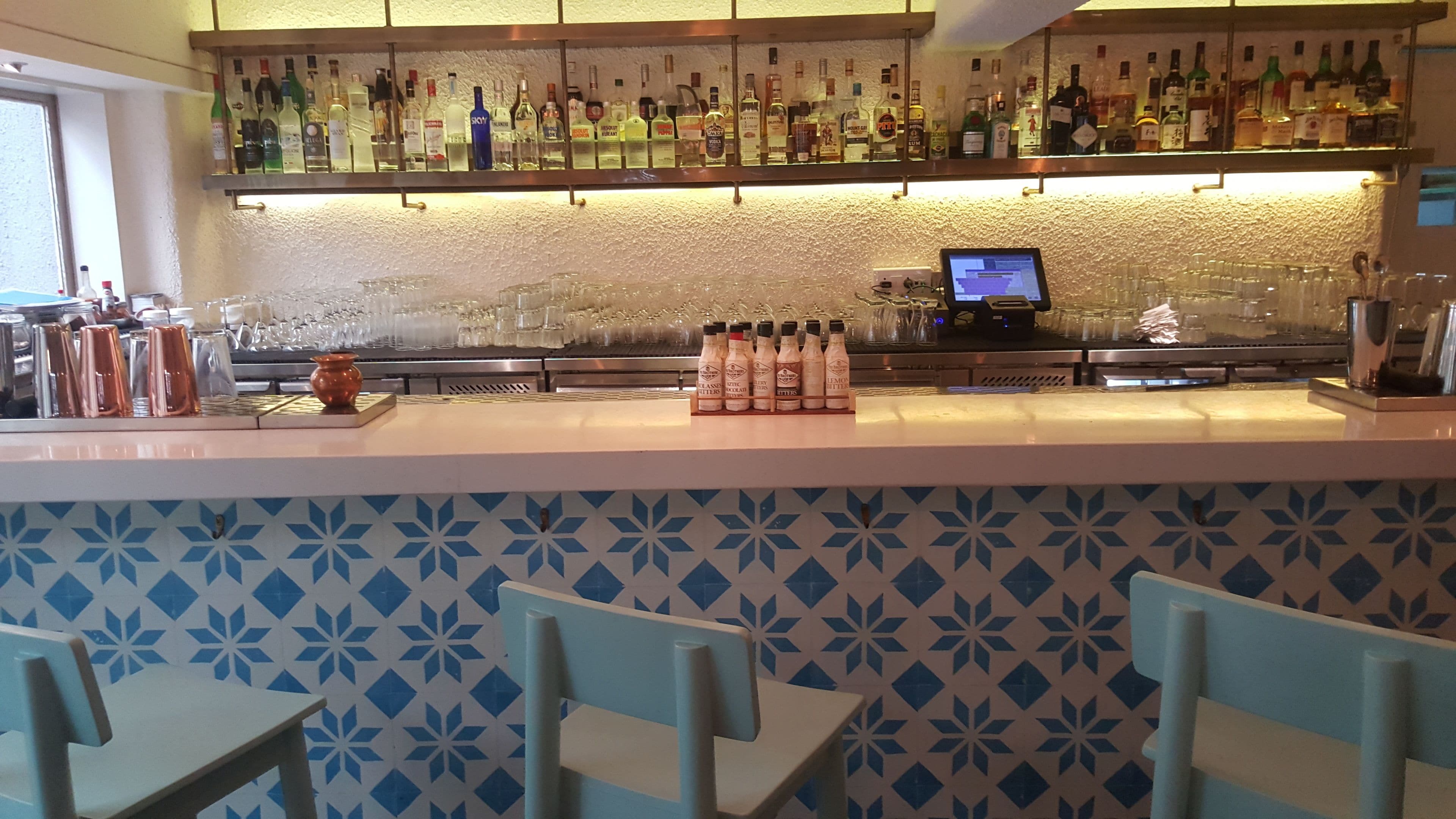 A bar area with a counter displaying various bottles and glasses, a digital payment terminal, and a patterned wall.