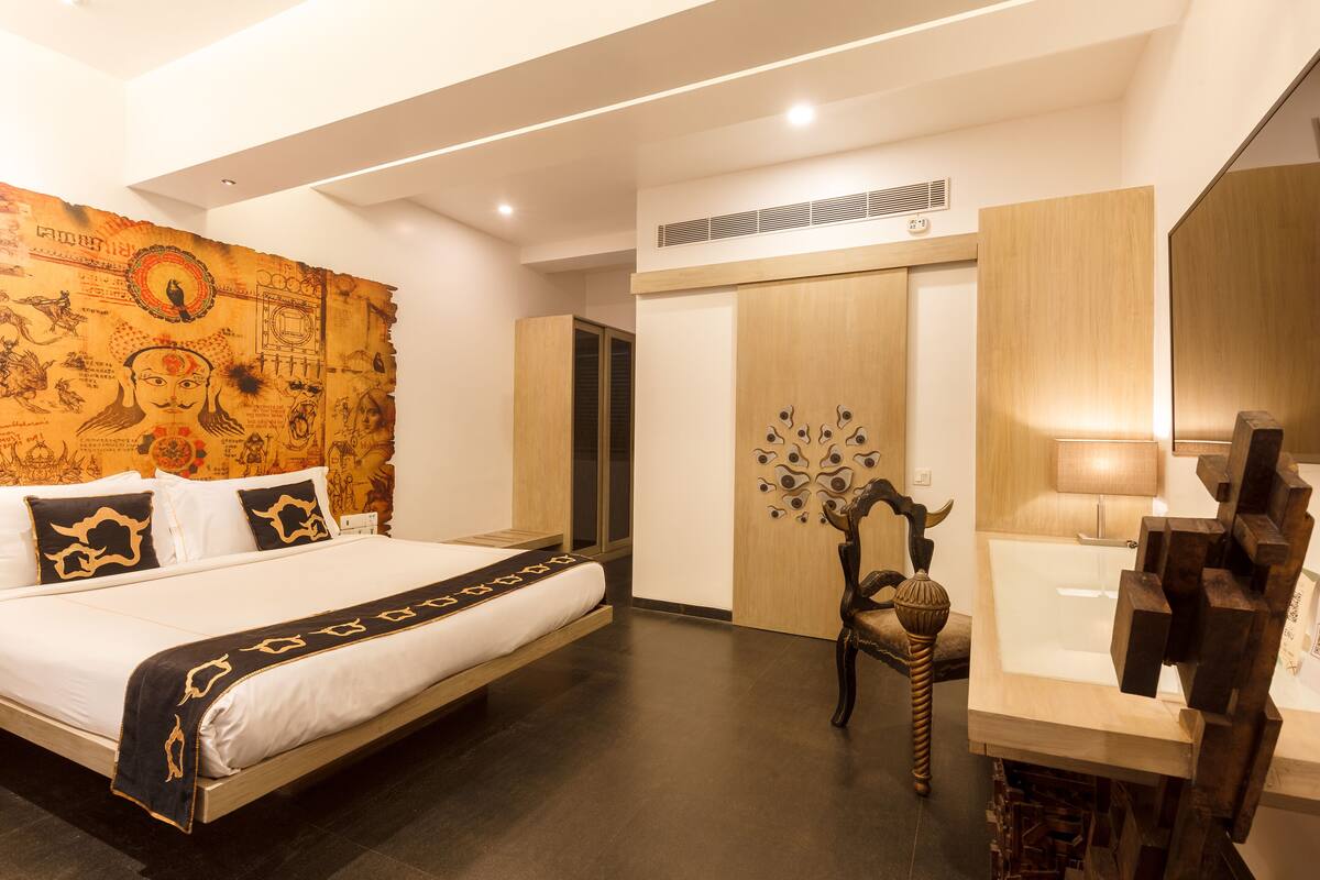 A modern hotel room with a bed, a wooden wardrobe, a bathroom with a sink and mirror, and a wall with a decorative tapestry.