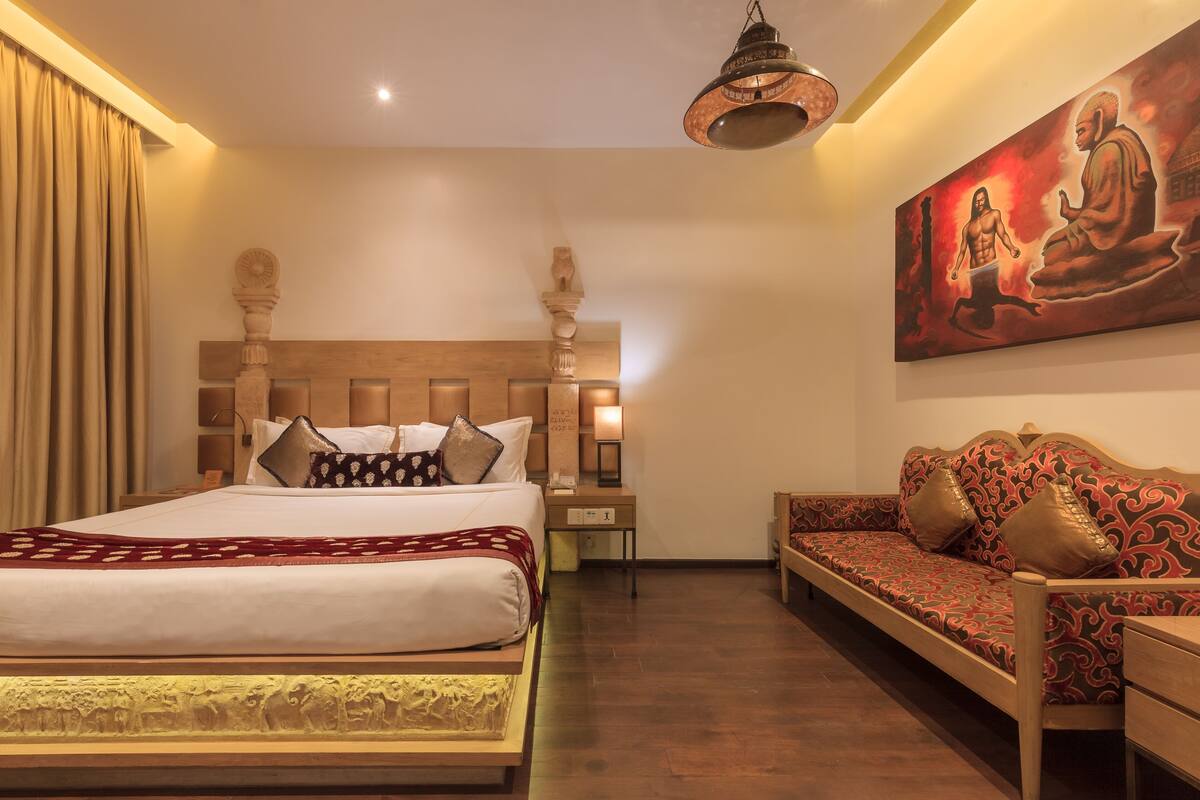 A hotel room with a large bed, a sofa, a nightstand, and a wall painting.