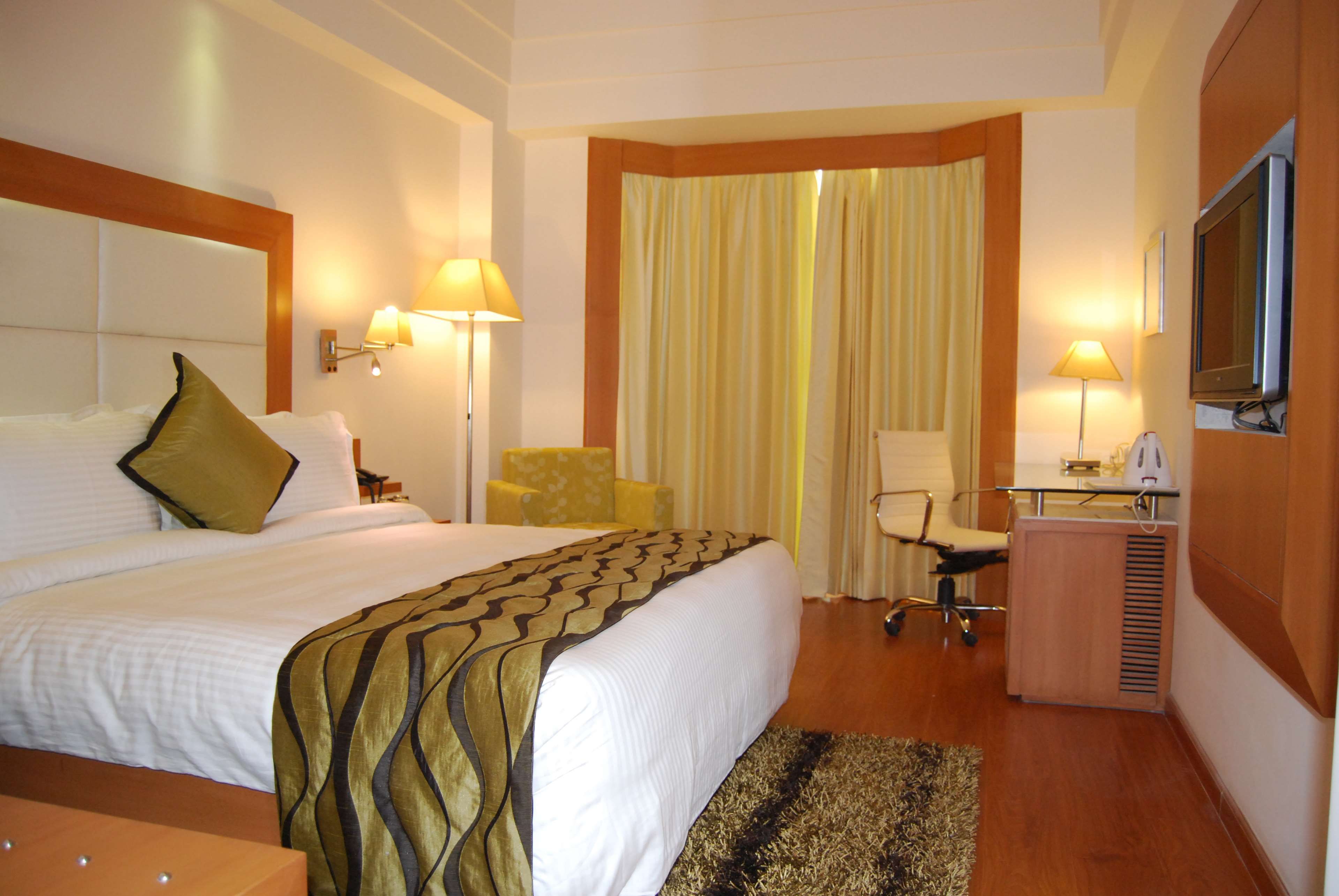 Ein Hotelzimmer mit einem großen Bett, zwei Nachttischlampen, einem Schreibtisch mit Stuhl, einem Fernseher und einem Fenster mit Vorhängen.