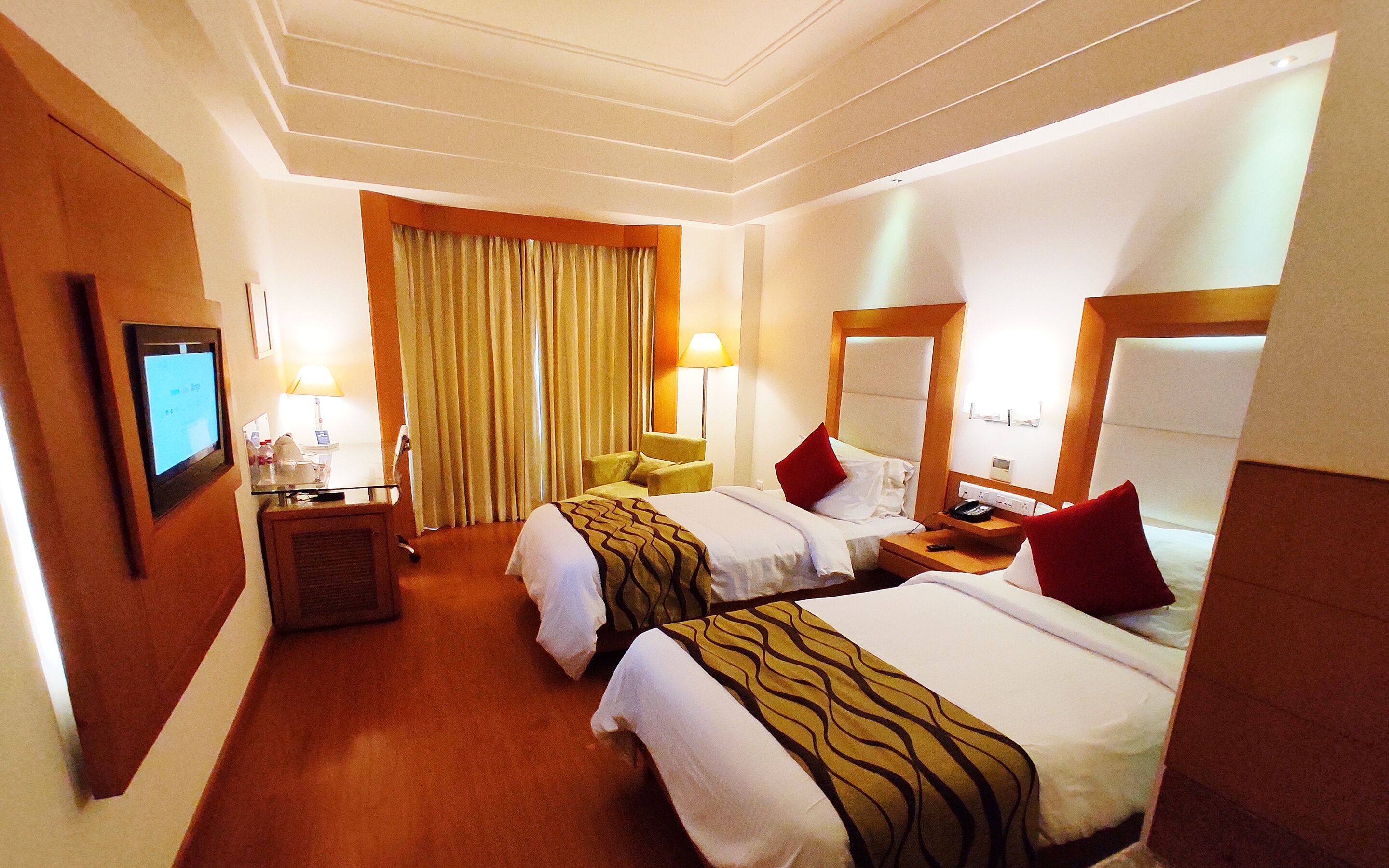 A hotel room with two beds, a desk, a chair, a TV, and a large window with curtains.