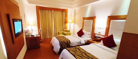 A hotel room with two beds, a desk, a chair, a TV, and a large window with curtains.