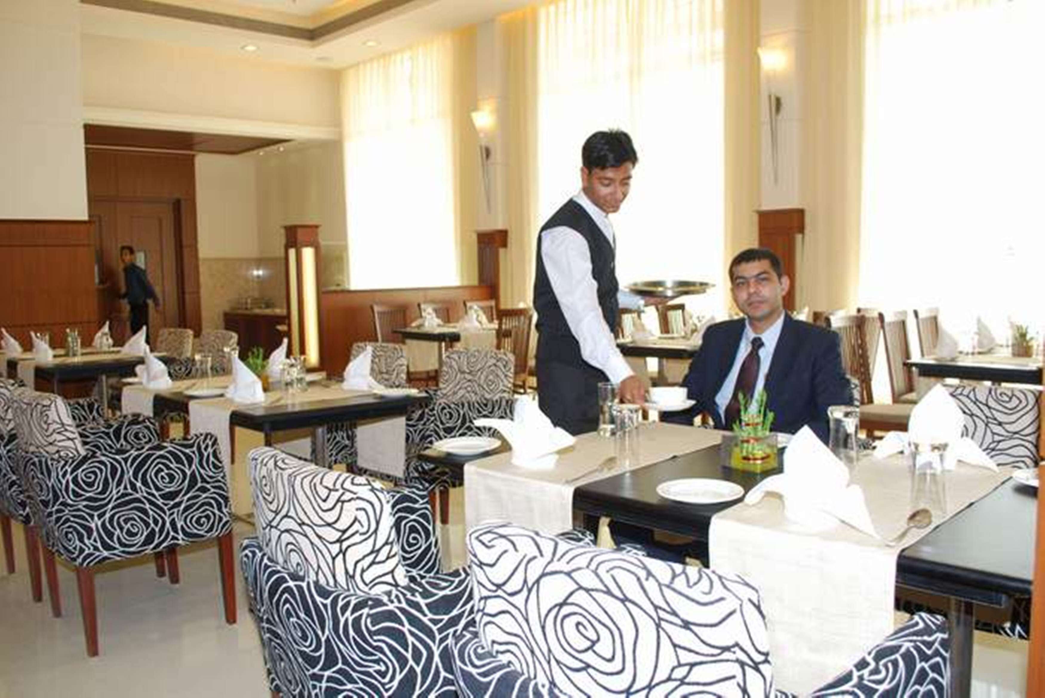 A restaurant with tables set for dining, a waiter attending to a guest, and a person in the background.