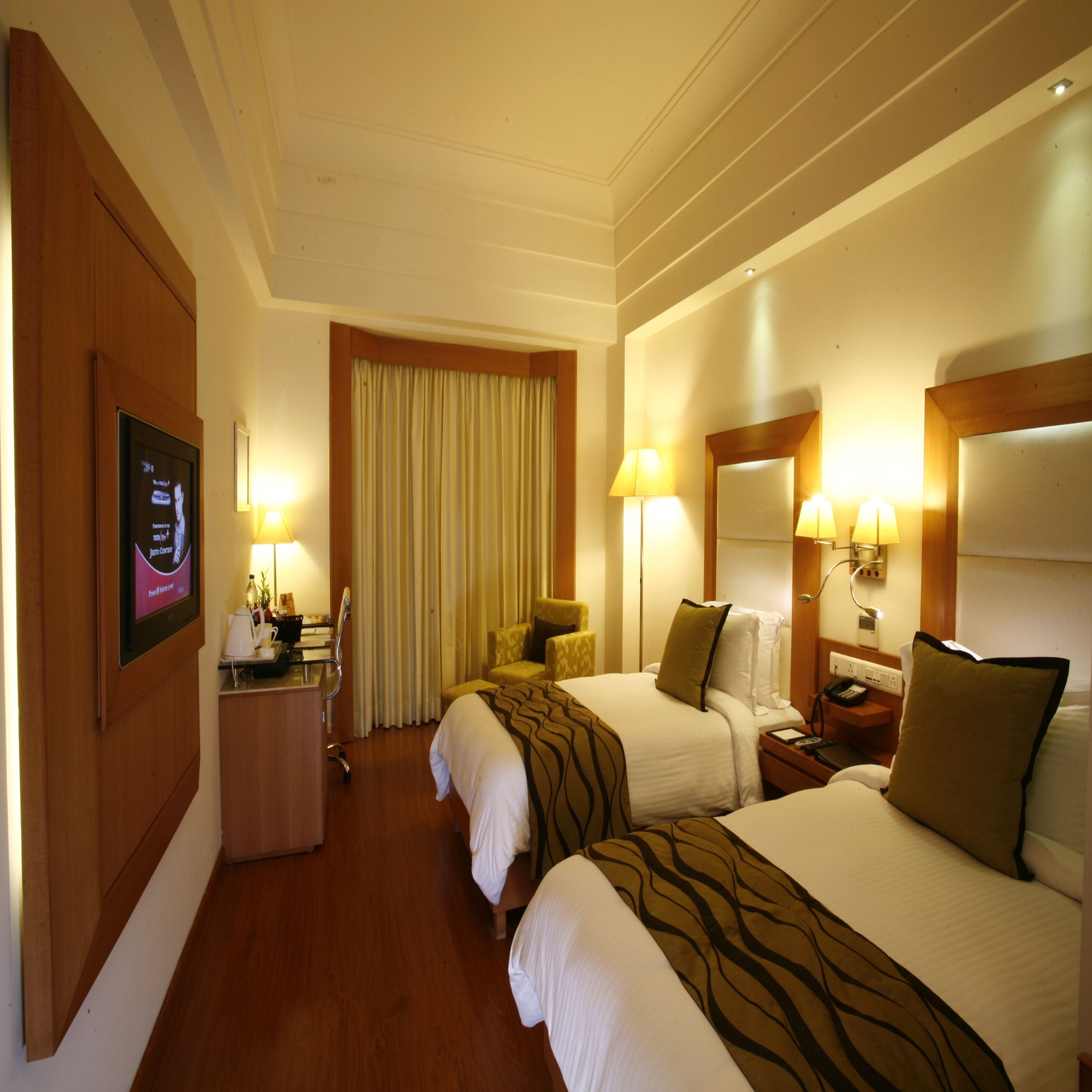 A hotel room with two beds, a desk with a computer, a chair, a lamp, and a TV mounted on the wall.