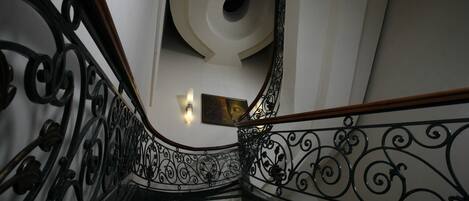 Una escalera con barandillas de metal negro ornamentado y un pasamanos de madera, que conduce a un rellano con una lámpara fijada a la pared y un cuadro enmarcado.