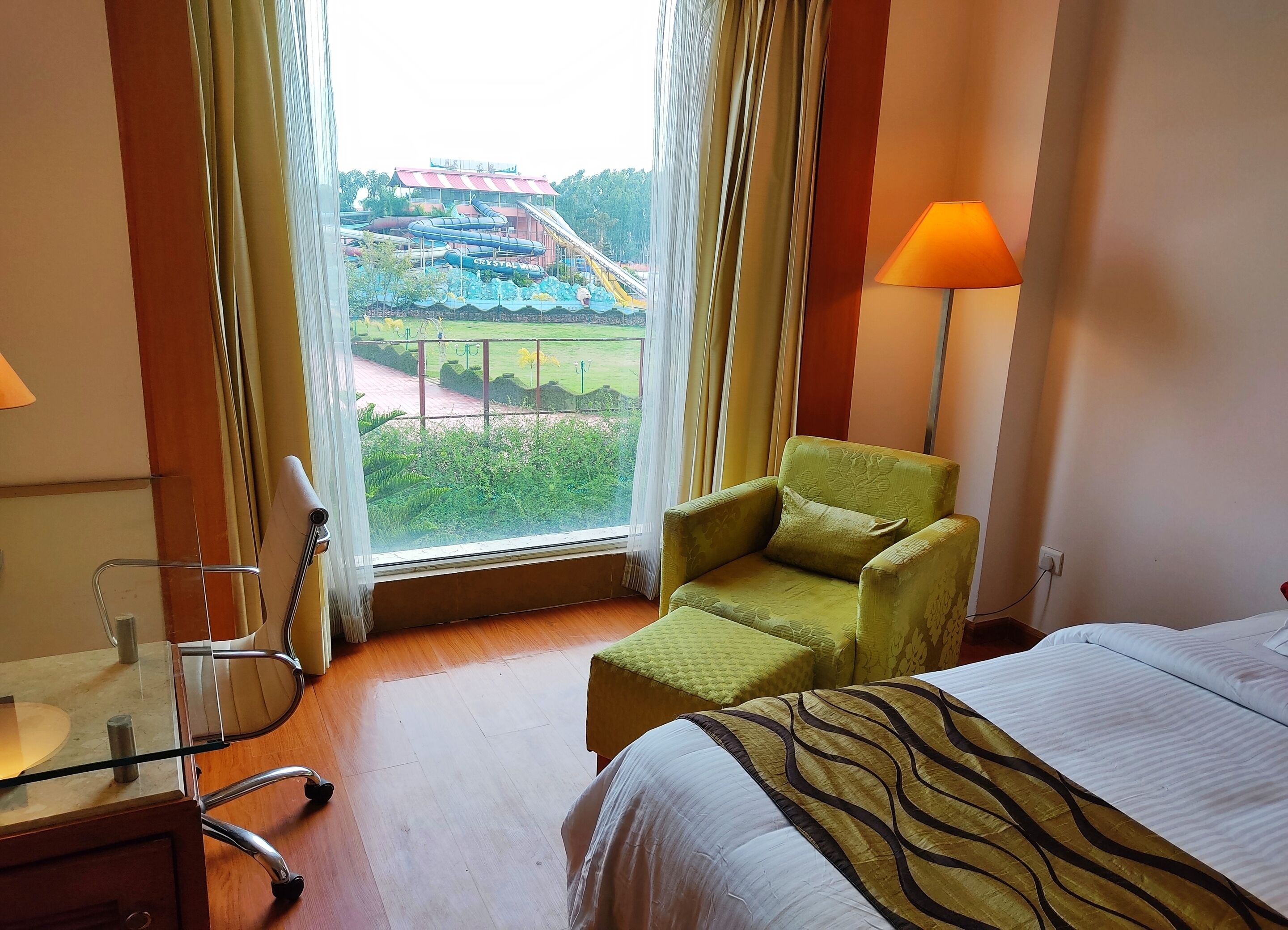 Habitación de hotel con una cama, un sillón verde, un escritorio con una silla y vistas a un parque acuático.