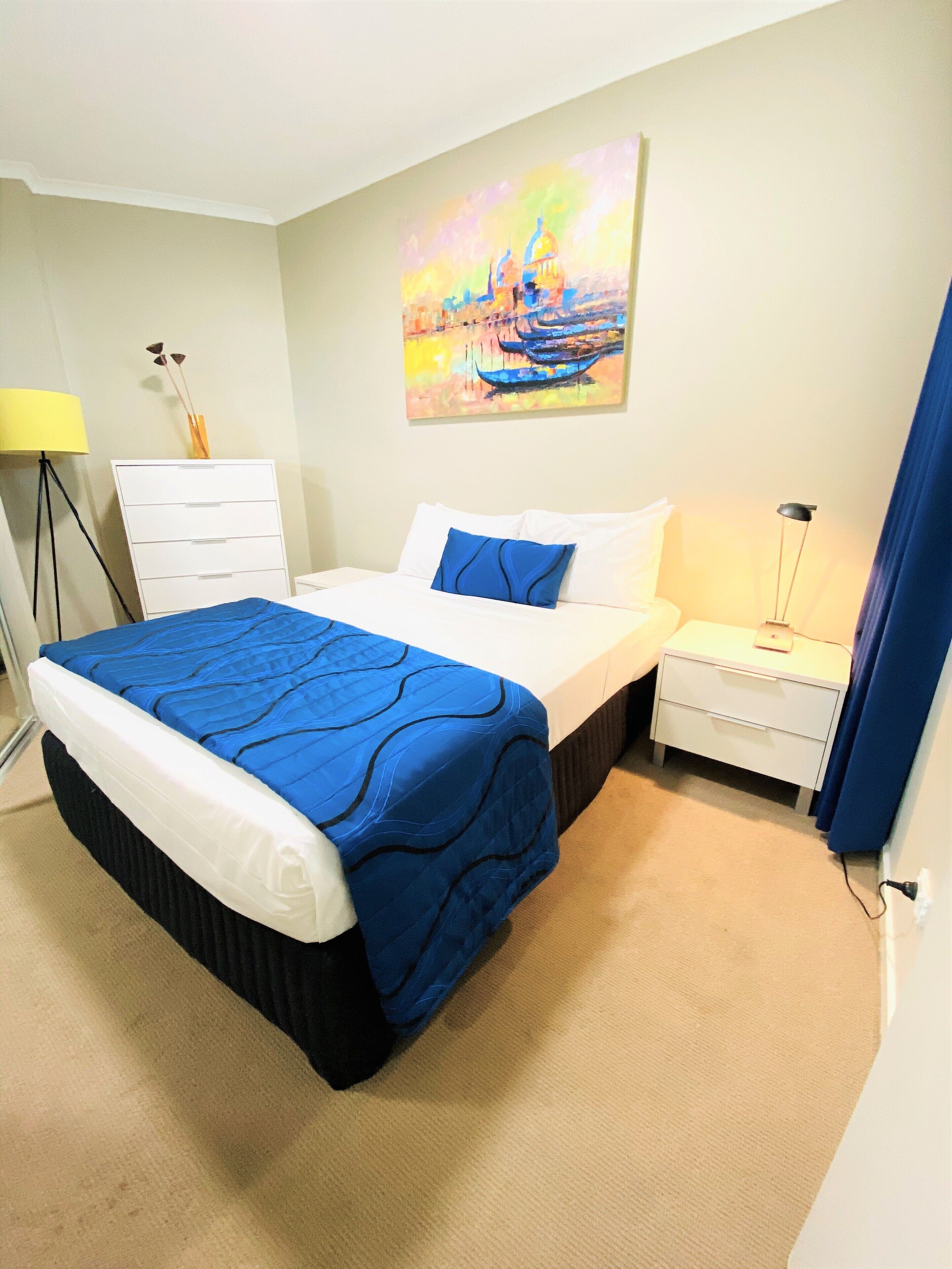 A hotel room with a bed, a nightstand, a lamp, a painting, and a dresser.