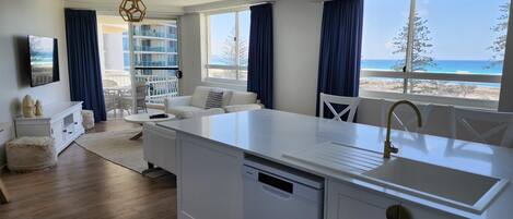 Una cucina moderna con lavastoviglie integrata, un piano di lavoro bianco e un lavello. C'è un'area pranzo con tavolo e sedie bianche, e vista sulla spiaggia tramite ampie finestre.
